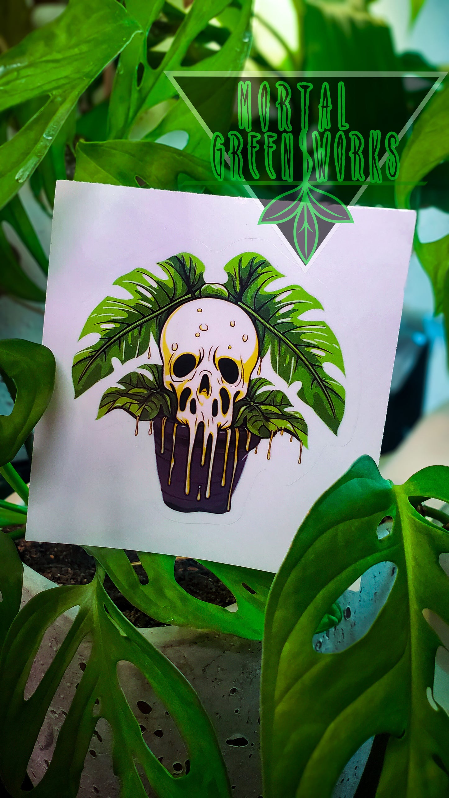 The sticker shows a skull with lush Monstera leaves sprouting from the top, set against a plant-filled background, branded with 'Mortal Greenworks'.