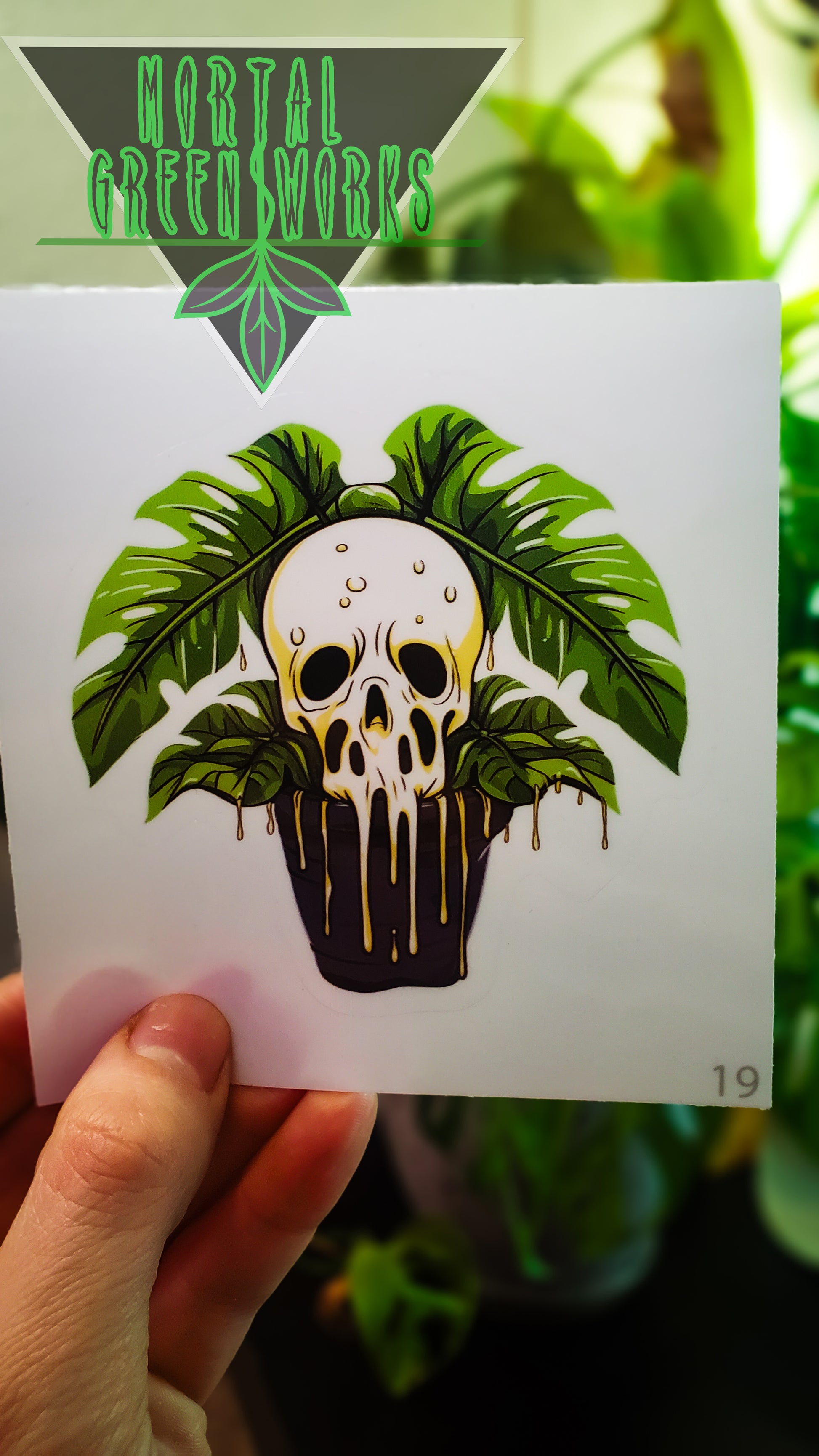 A hand holds a sticker of a skull with Monstera leaves as hair and dripping plant matter, with 'Mortal Greenworks' logo above.