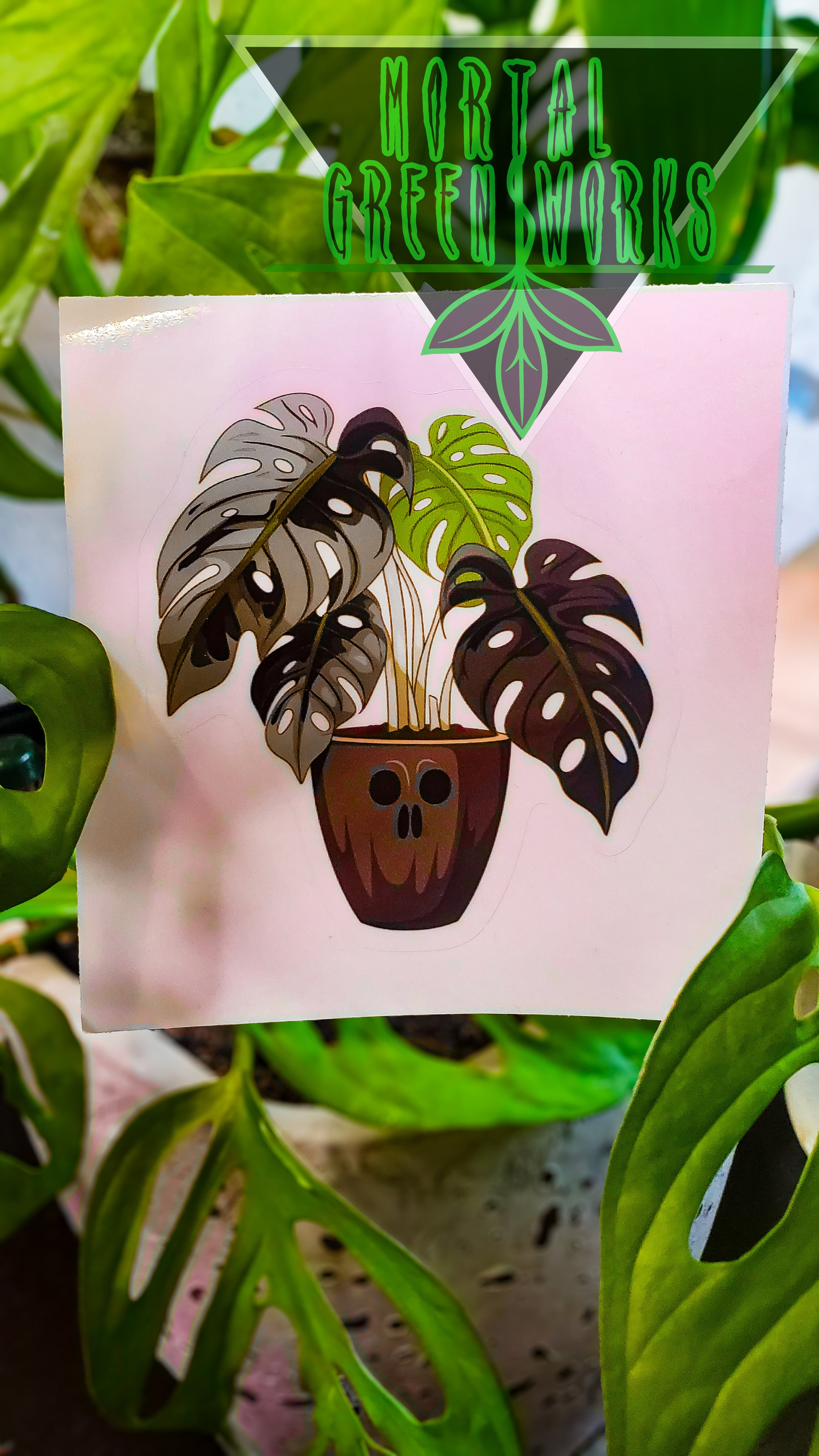 A sticker of a monstera in a skull pot in front of live greenery with the 'Mortal Greenworks' logo overlay.