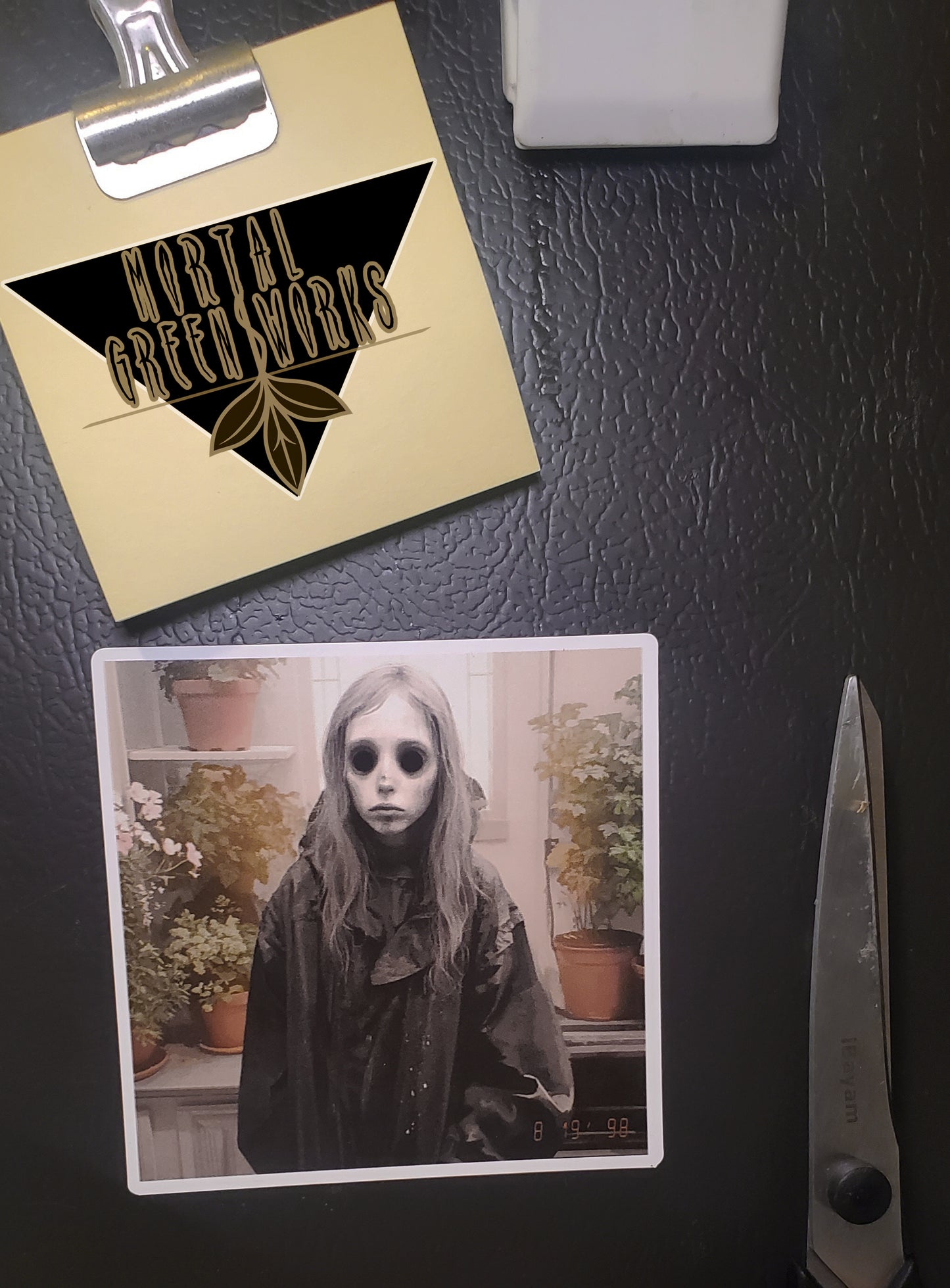 A fridge adorned with a magnet of a ghostly, pale child in a sunroom, combining nostalgia with a chilling twist, marked by Mortal Greenworks.