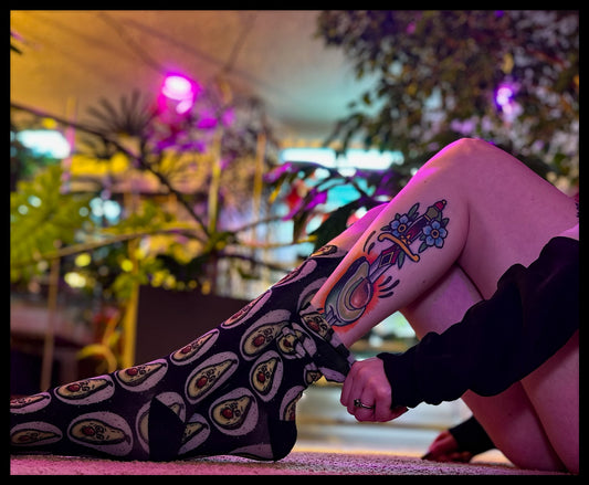 Person in a moody indoor garden setting wearing avocado-print socks, highlighting a bold tattoo, perfect for a trendy and artsy aesthetic