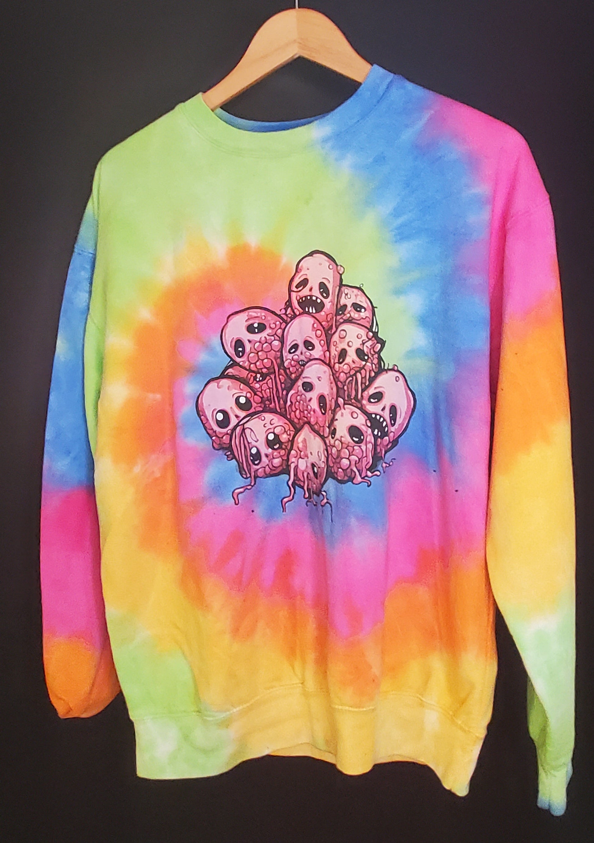 Hanging against a dark background is a tie-dye sweatshirt with a vivid color palette including yellow, green, blue, and pink. At the center of the sweatshirt is a graphic of a group of cartoon raspberries with expressive faces. The sweatshirt has a crew neck and long sleeves with a relaxed fit, showcasing the bright, eye-catching tie-dye pattern.