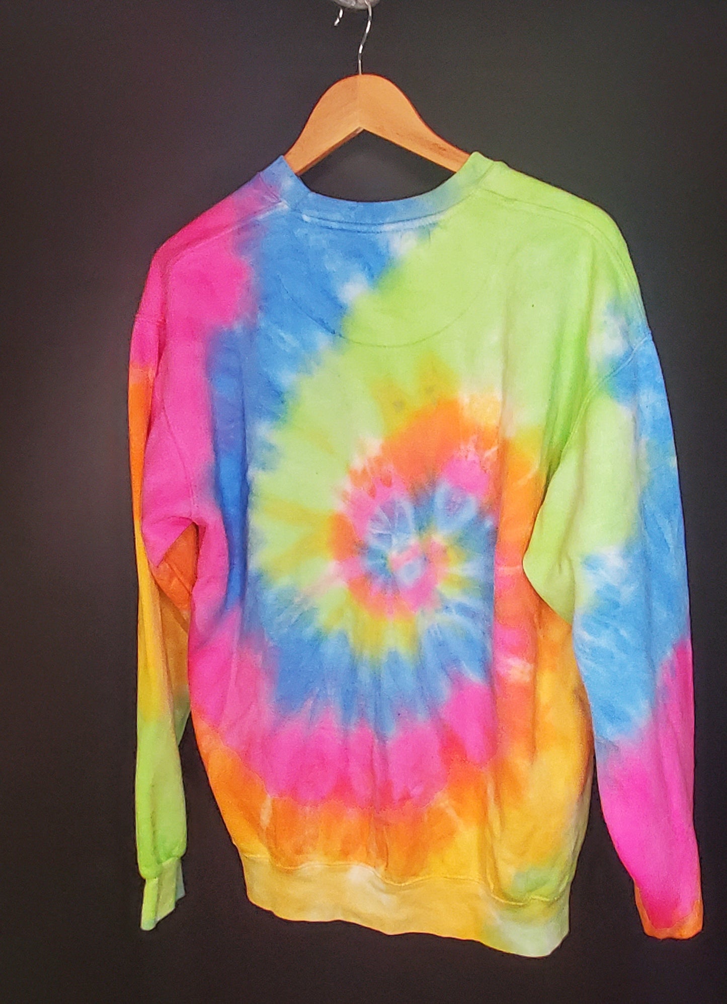 A tie-dye sweatshirt is displayed on a wooden hanger against a dark background. The sweatshirt features a swirl of vibrant colors, including yellow, green, blue, and pink, creating a radiating pattern from the center. The sweatshirt has long sleeves and a crew neckline, with the dye pattern giving it a playful and retro look