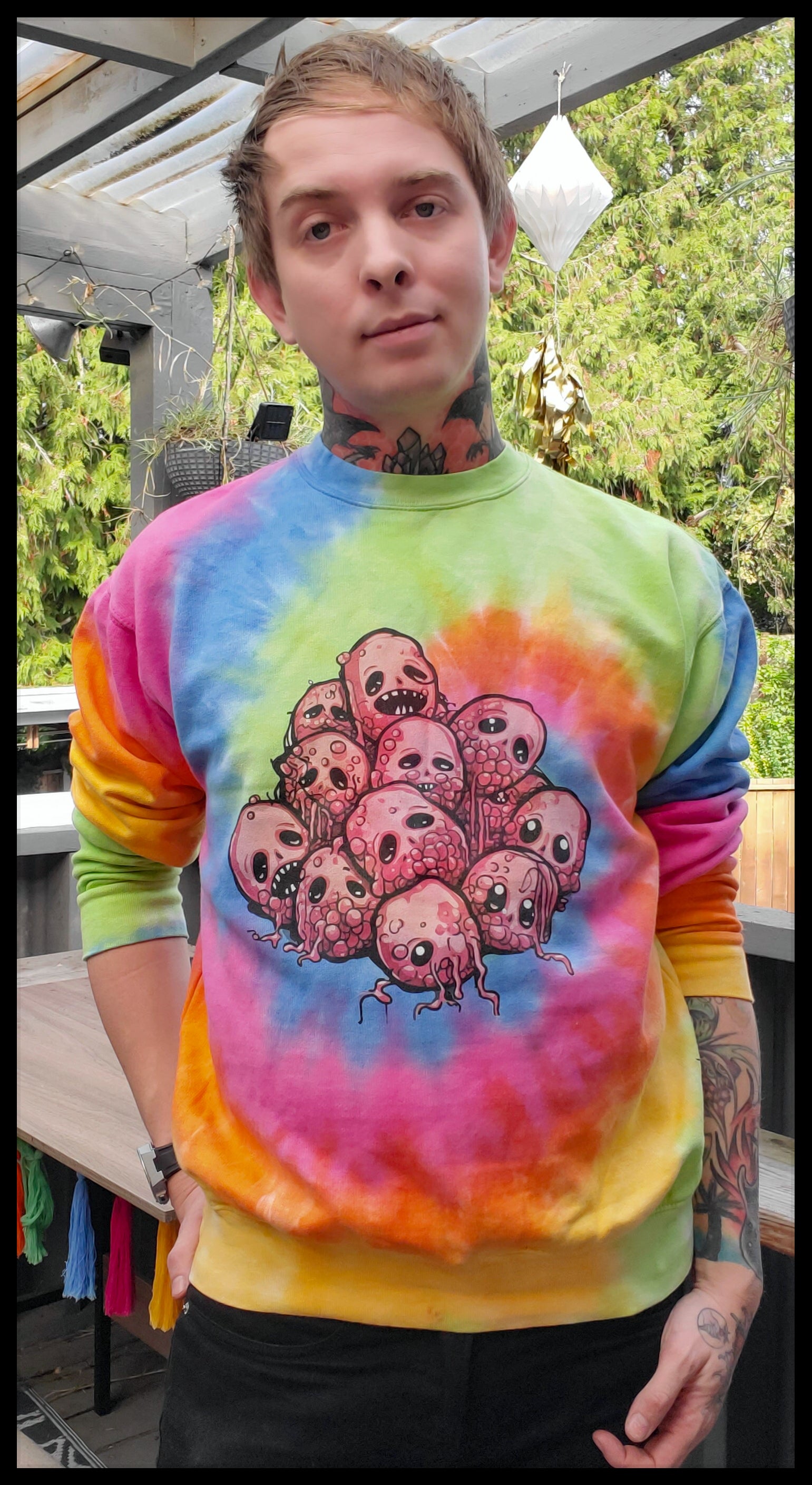 The individual is wearing a vibrant tie-dye sweatshirt with a central graphic featuring a group of cartoon raspberries with various expressions. The colors of the sweatshirt transition from yellow at the bottom to green, blue, and pink, with the sleeves ending in a deep orange. The person has short hair, visible tattoos, and is wearing a smartwatch on the left wrist, with black pants. They are standing on an outdoor deck with plants and decorative items in the background.