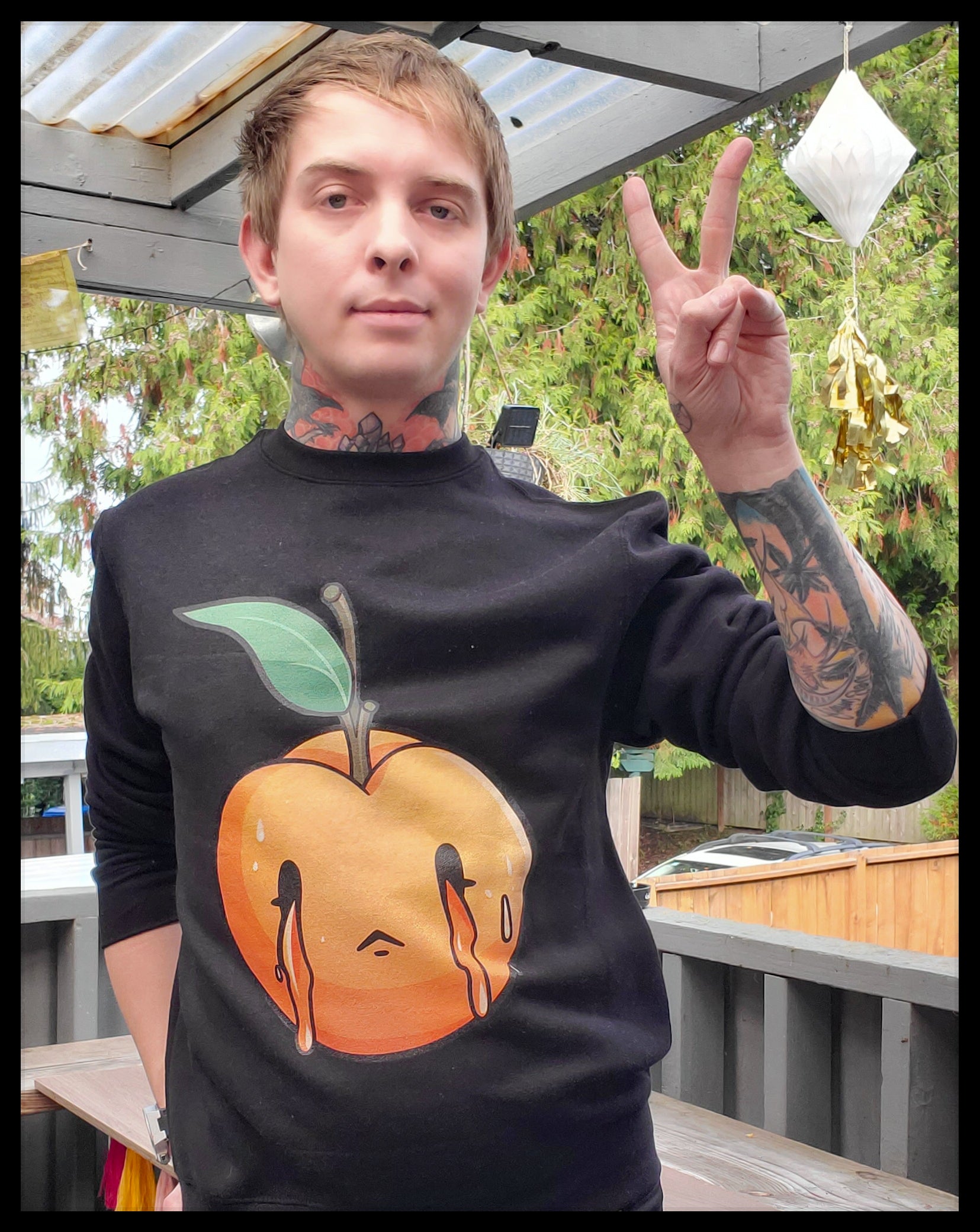 A person is posing on a deck, making a peace sign with their right hand, wearing a black sweatshirt. The sweatshirt showcases a central graphic of a peach with a sorrowful face, with two tears rolling down. The peach is topped with a green leaf and stem. The person's tattoos are prominently displayed on their arms and neck, and the outdoor setting includes potted plants and translucent hanging decorations.