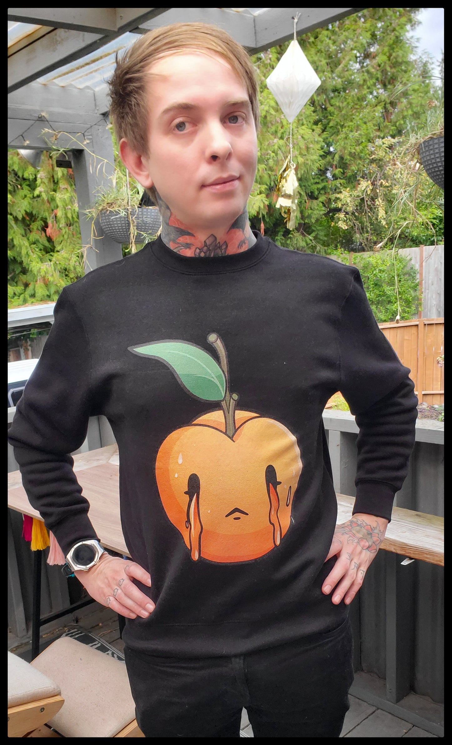 A person stands outdoors wearing a black sweatshirt with a large, vivid graphic of a sad, crying peach on the front. The peach is depicted with a teardrop design and a melancholic expression. The sweatshirt has a round neck and long sleeves, fitted cuffs, and a relaxed silhouette. The individual pairs the sweatshirt with black pants and sports a smartwatch on the wrist, with tattoos visible on the hands and neck, set against a backdrop of a deck and greenery.