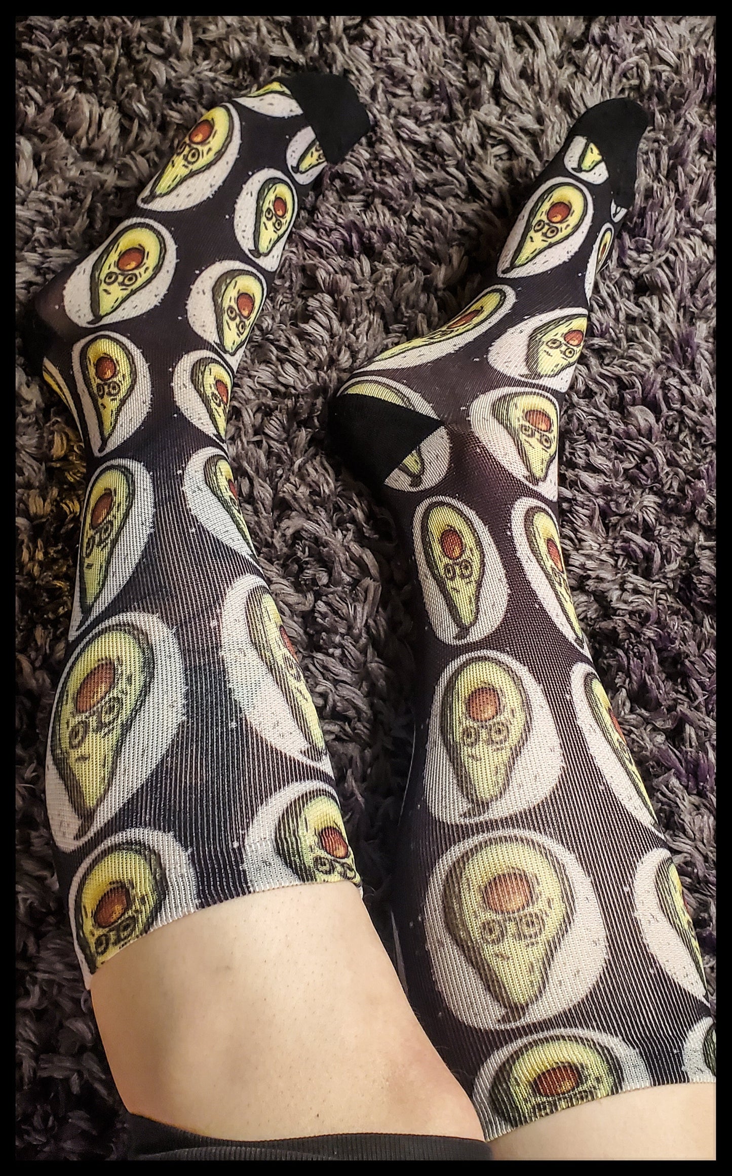 A pair of black socks stretched over the wearer's legs, featuring a pattern of avocados with a brown pit stuffed in the mouth, giving a look of surprise, and two round eyes. The avocados are set against a white painted circular background, accentuating the design on the black fabric of the socks. The image showcases the socks against a textured grey carpet, emphasizing the unique and playful avocado motif.