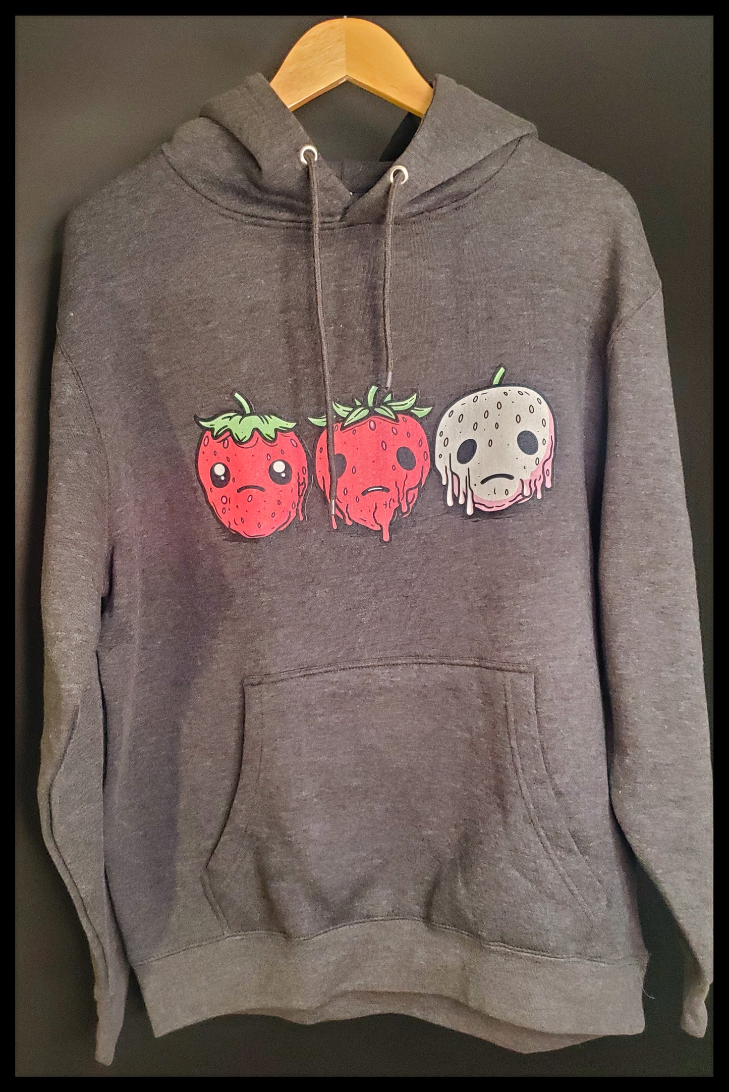 Gray hoodie with three sad strawberries designed on the front, sits on a wooden hanger against a dark background