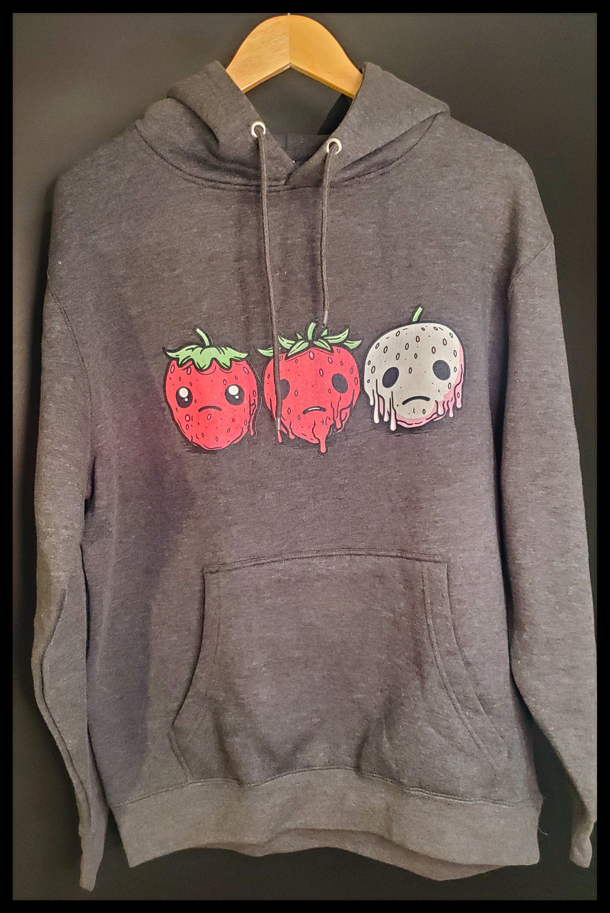 Gray hoodie with three sad strawberries designed on the front, sits on a wooden hanger against a dark background