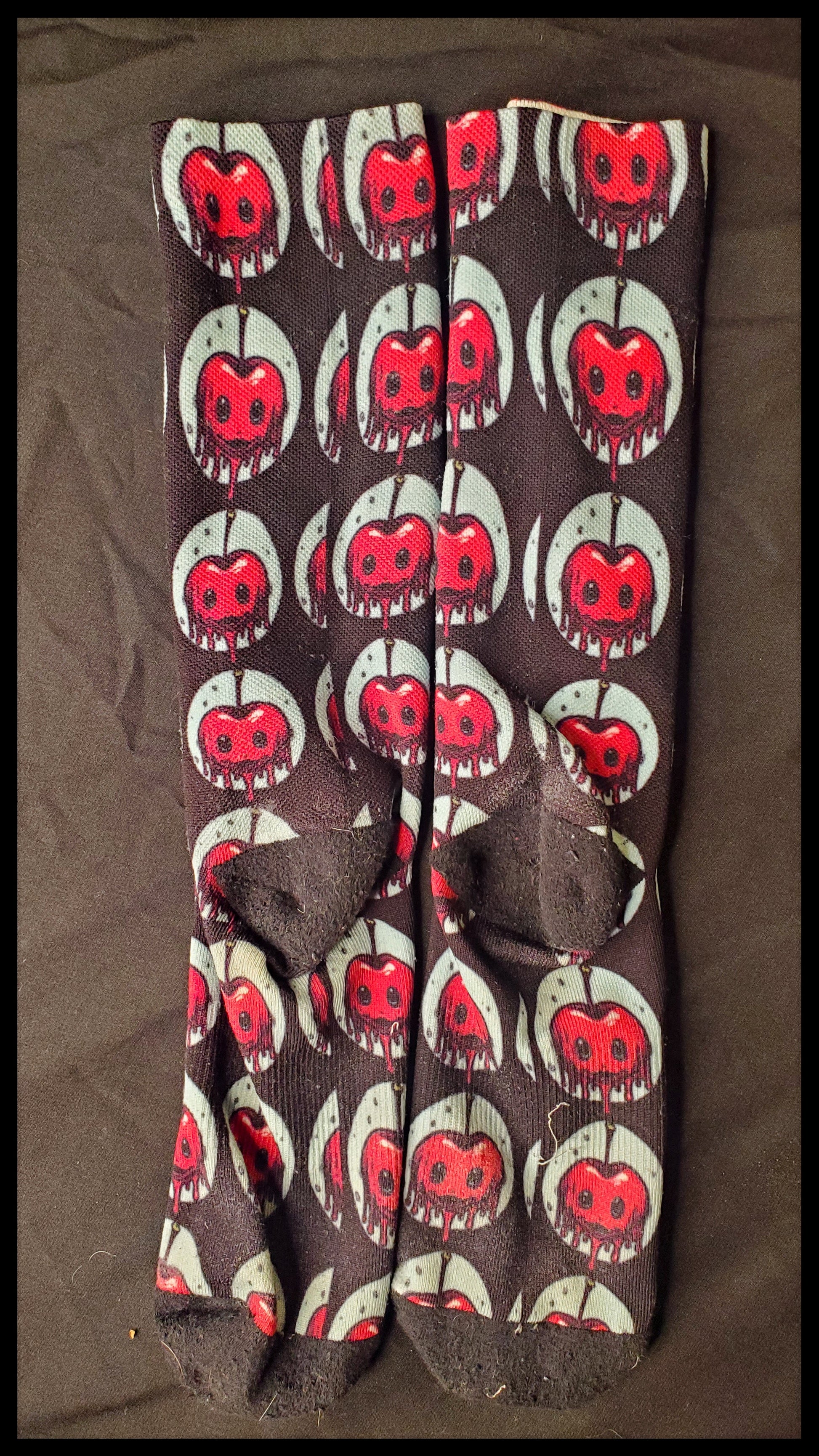 A pair of flat-laid socks with a horror-themed pattern of red apples with white highlights and black facial features, mimicking a spooky expression. Each apple is depicted with a dripping blood effect. The socks are black with a ribbed texture, featuring a black heel, toe, and cuff. The background is a solid darker shade, providing contrast to the socks' pattern.