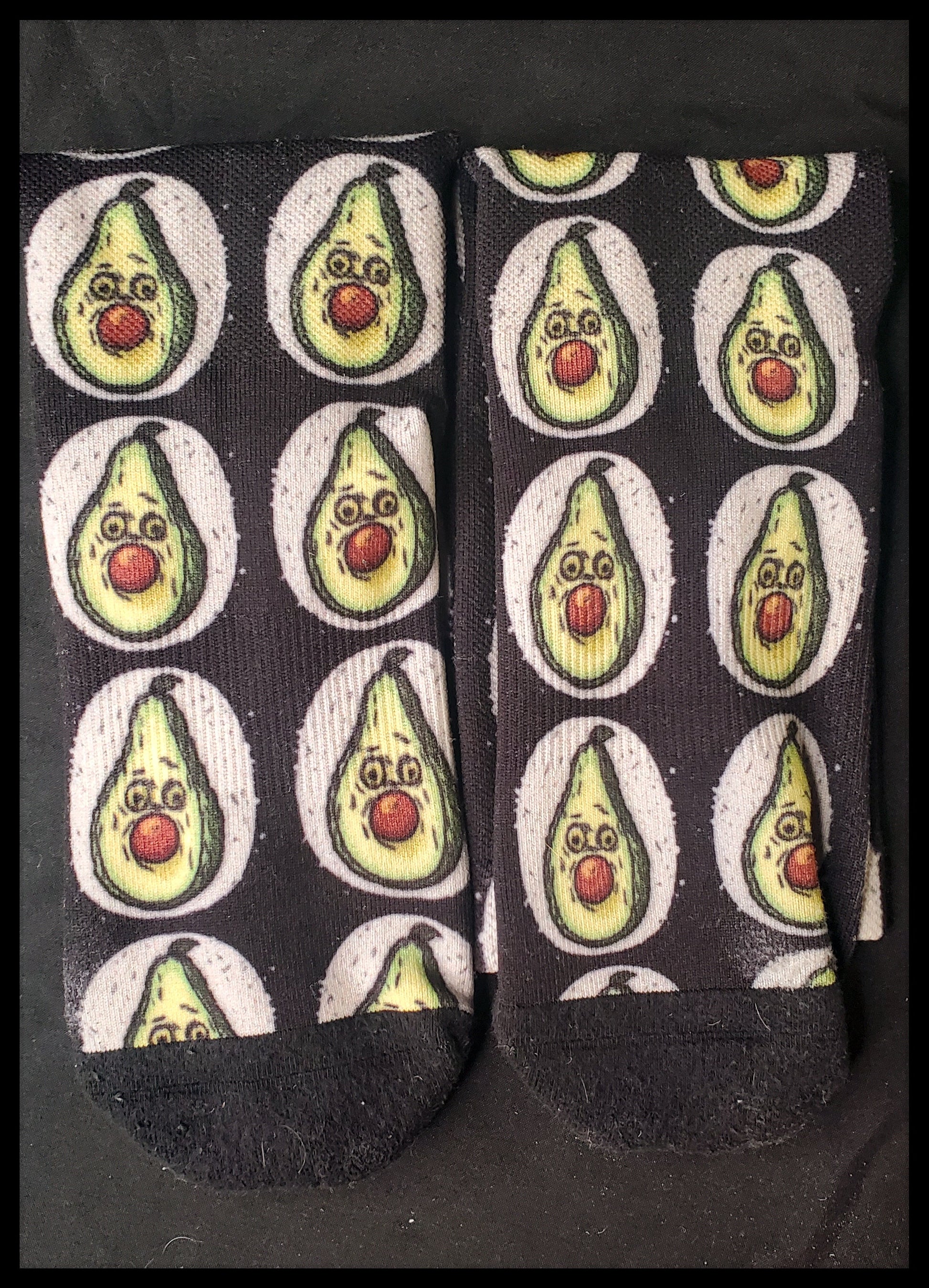 A pair of black socks laid flat, displaying a playful avocado pattern with each avocado showing a facial expression where the brown pit serves as a stuffed mouth. The avocados are depicted with green skin and two round, surprised eyes, set against a white circular background for contrast. The socks have black heels, toes, and cuffs, and the image captures the whimsical design well