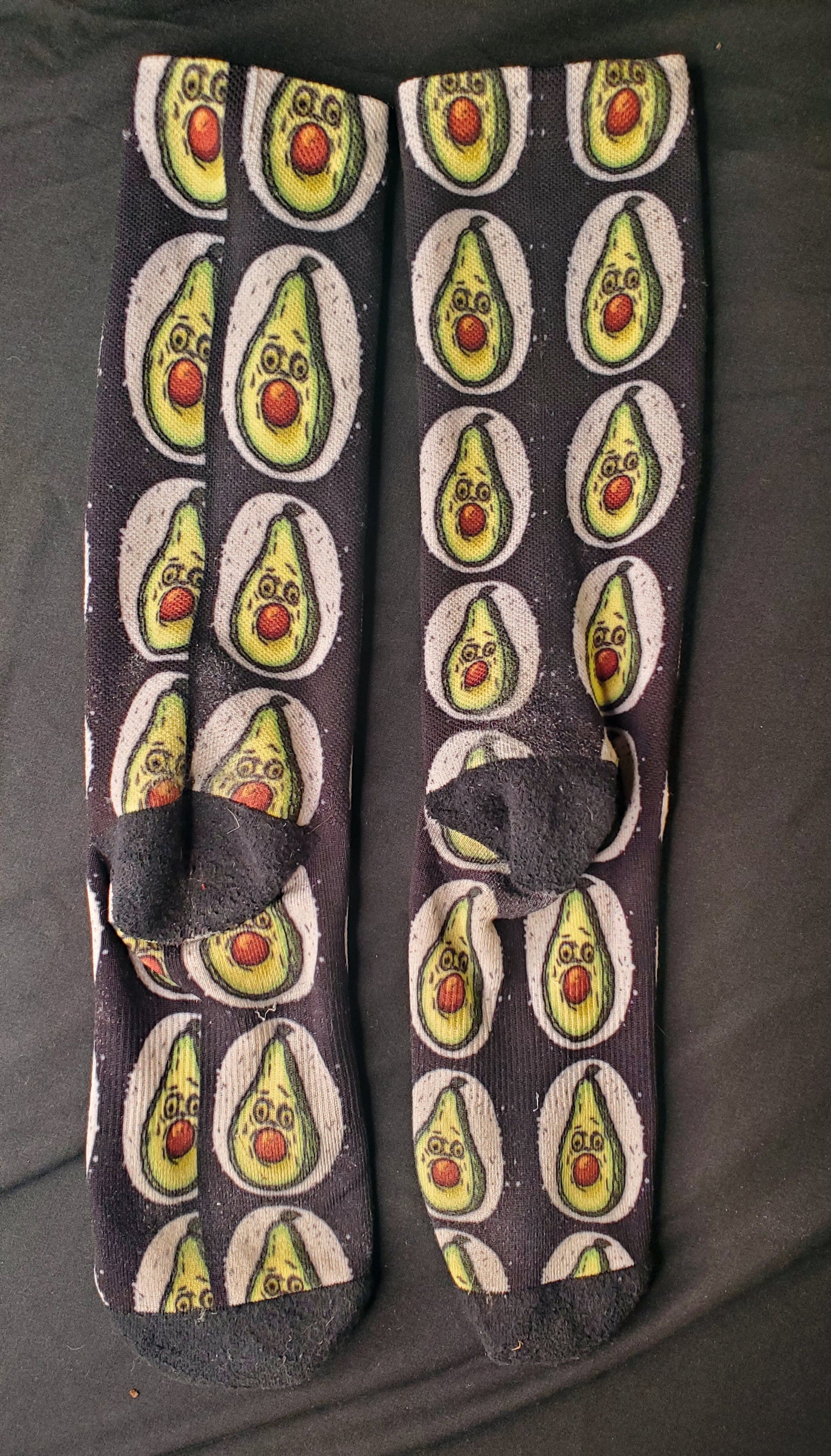 A pair of black socks displayed flat, featuring a pattern of avocados with surprised expressions, the brown pit positioned as if it's stuffed in the mouth, and two round eyes. Each avocado is set within an off-white painted circle against the black sock fabric, creating a striking contrast. The design adds a whimsical and playful element to the socks.