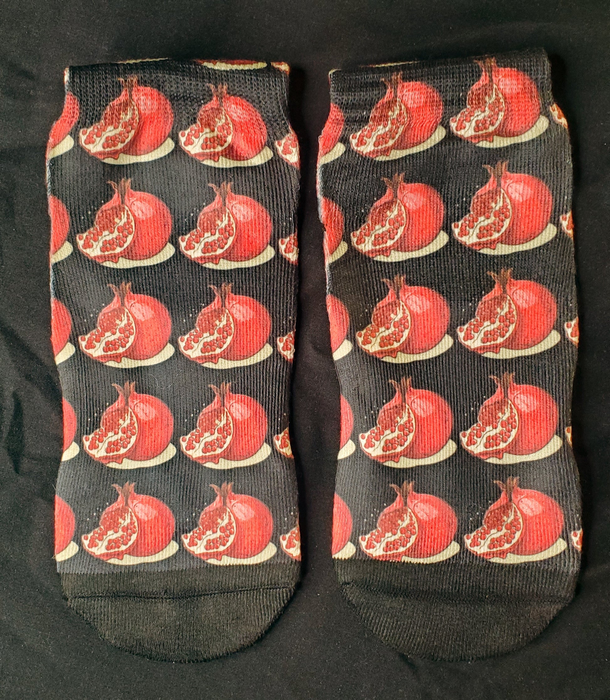 The socks are adorned with a vivid pattern of red pomegranates against a contrasting dark backdrop, each fruit detailed with highlighted seeds and a subtle sheen, infusing a pop of color and a touch of playful elegance to the design.