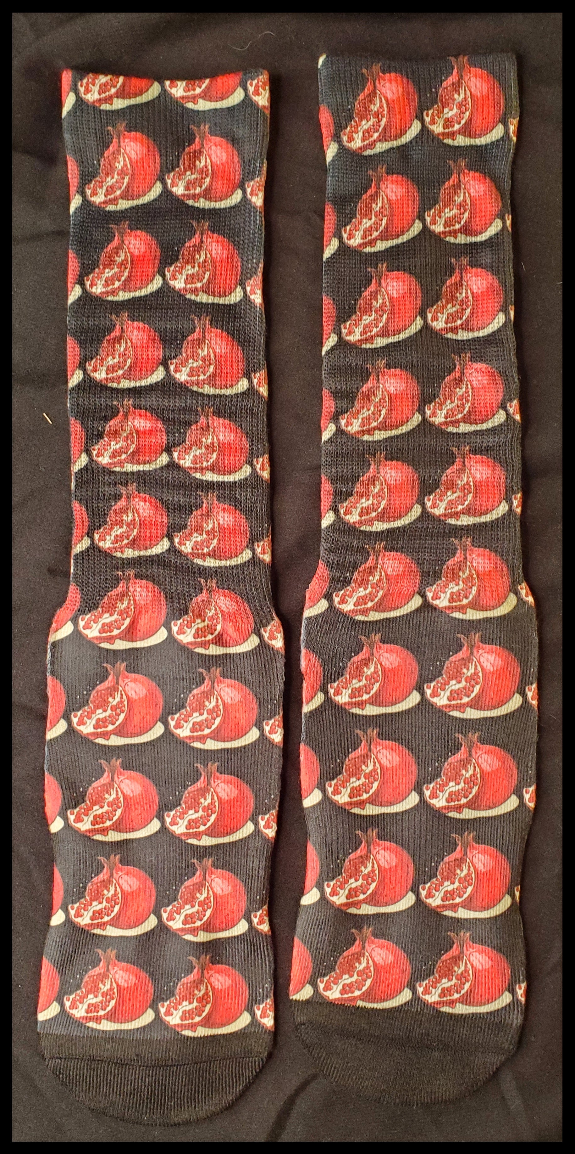 The socks feature a pattern of red pomegranates with highlighted seeds, set against a dark backdrop. The vivid contrast accentuates the fruit's lush appearance.