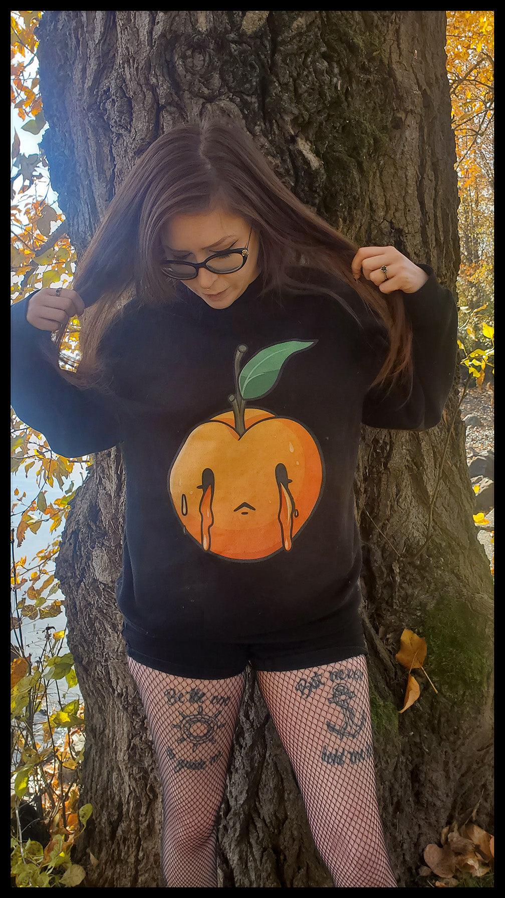 A girl is posing with a hoodie adorned with an artistic design of a peach with a tearful face, set against a dark background that accentuates the vivid colors of the peach illustration. The design conveys a sense of emotional depth