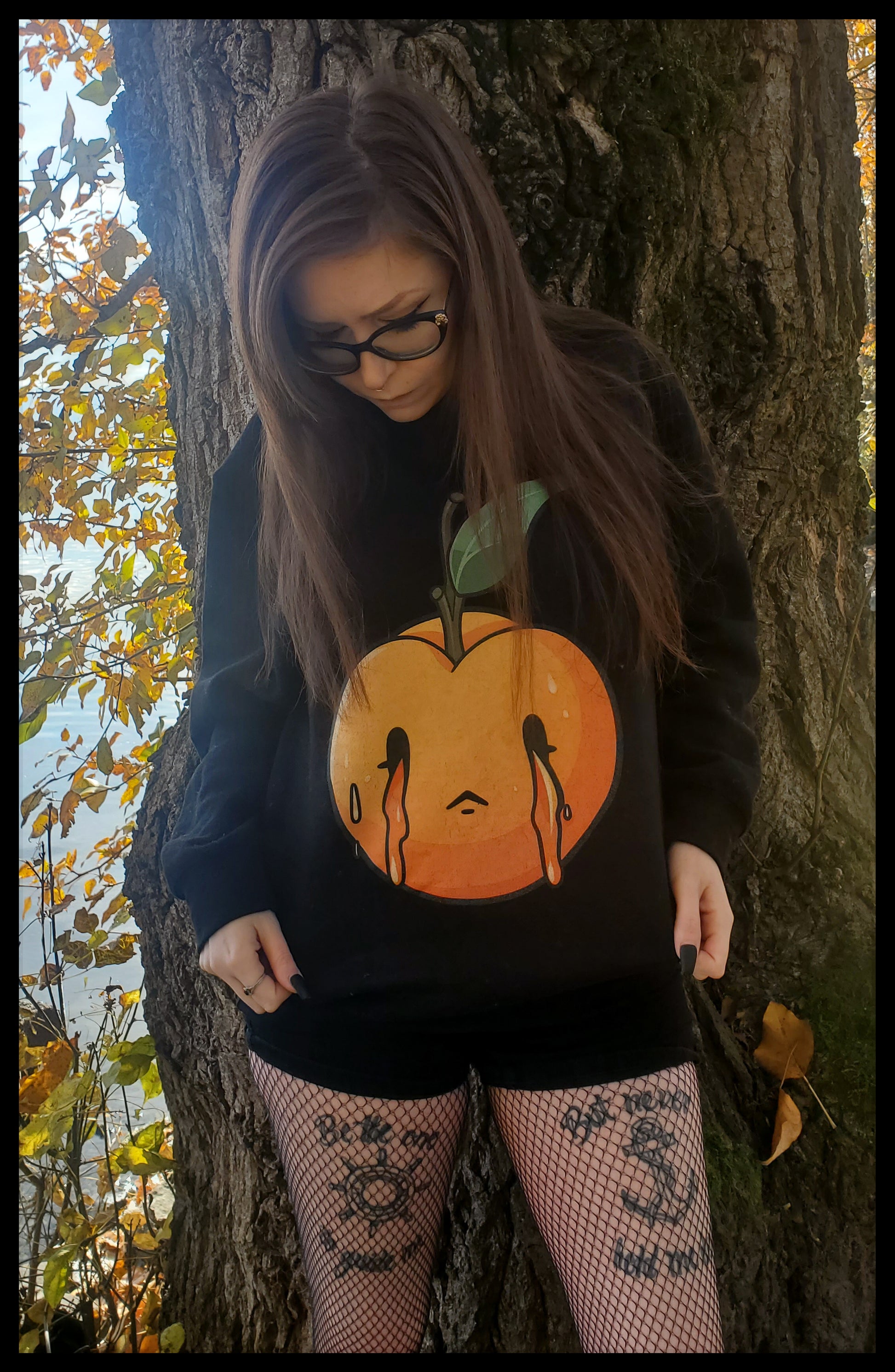 The person is wearing a sweatshirt featuring a stylized peach with a melancholic expression, showcased against the sweatshirt's dark fabric. The peach's vivid orange hue and the teardrops add a unique, emotional character to the design.