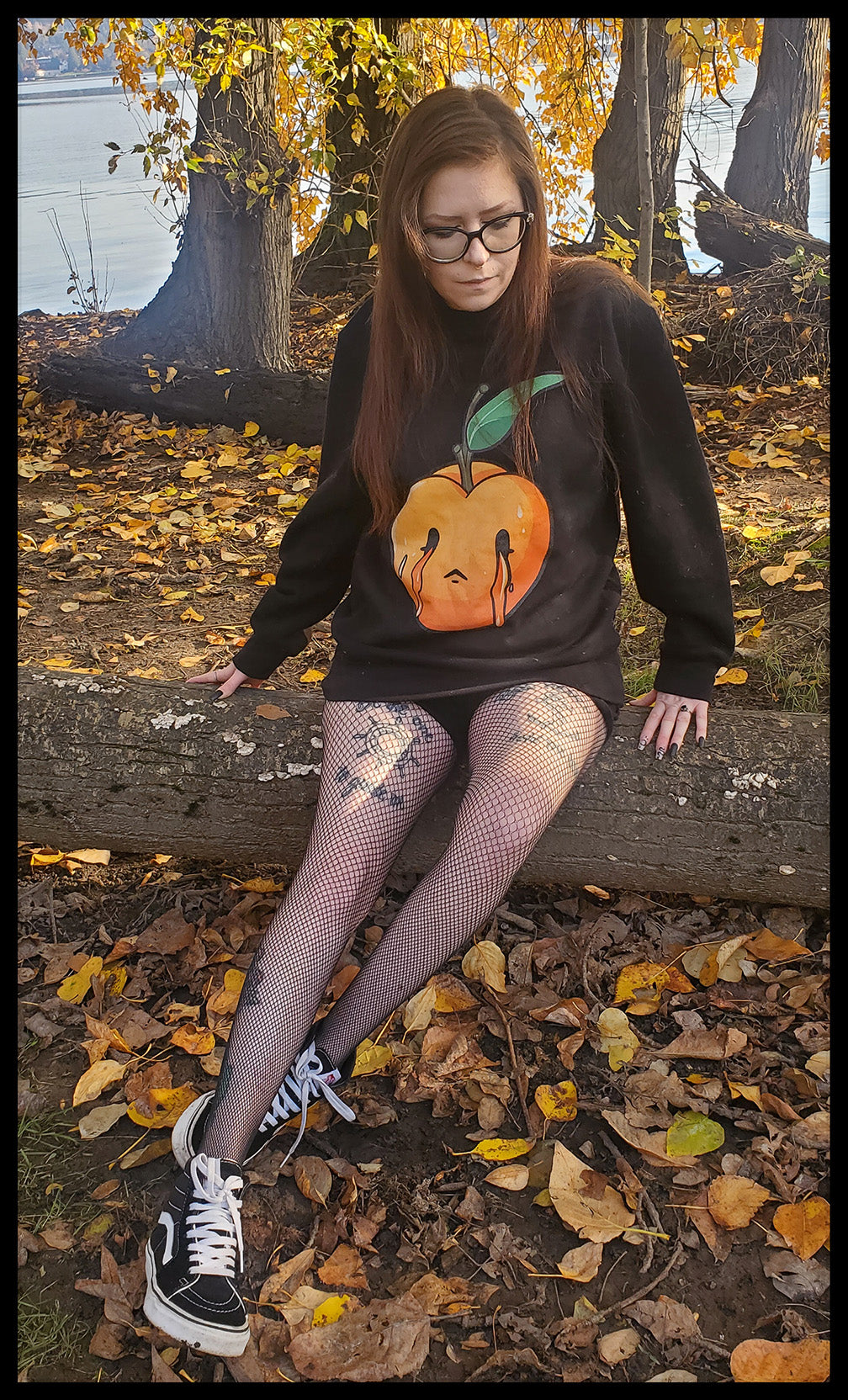 A girl is seated outdoors wearing a black sweatshirt featuring a large, anthropomorphized peach design with a tearful expression, set against a backdrop of fallen autumn leaves. The peach illustration is central on the garment