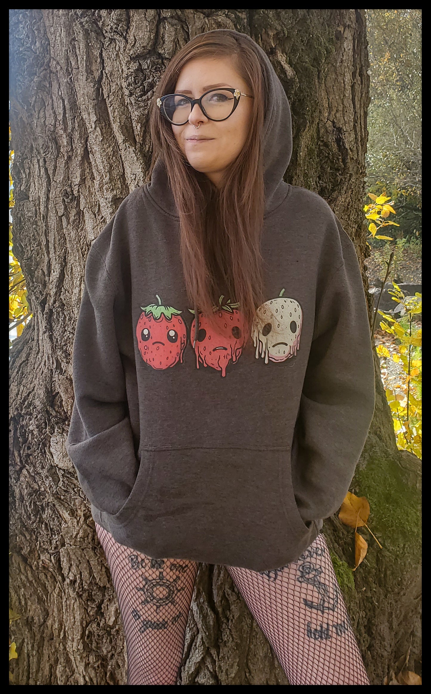 a person leaning against a tree, wearing a dark hoodie with a horror-themed fruit design on the front. They are wearing glasses and fishnet tights with tattoo designs visible on their legs. The background suggests an outdoor setting with trees and yellow leaves