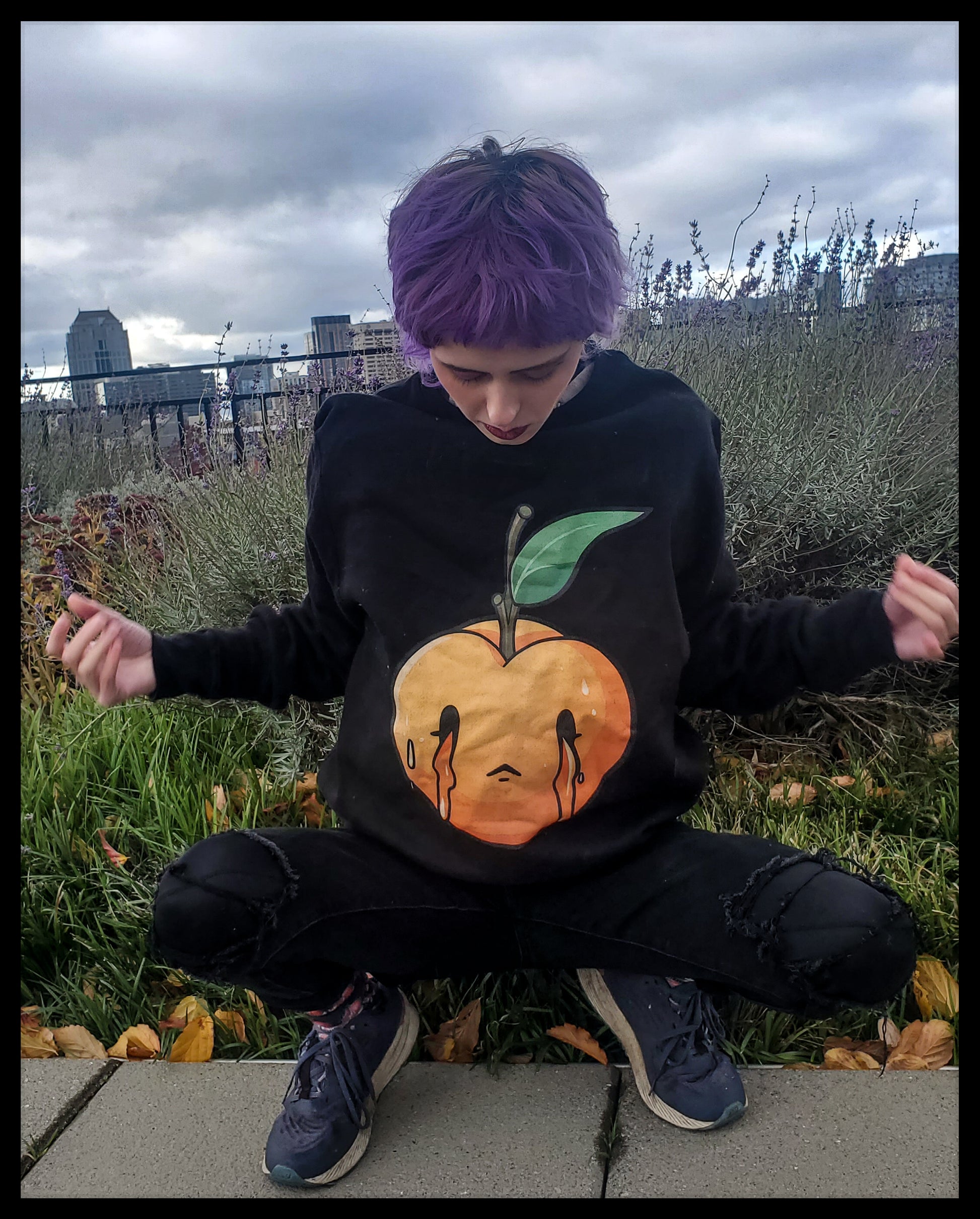 Person with short purple hair, meditating in a squatting position. They are wearing a black sweatshirt adorned with a stylized peach design, complemented by black torn jeans and sneakers, set against an urban garden backdrop