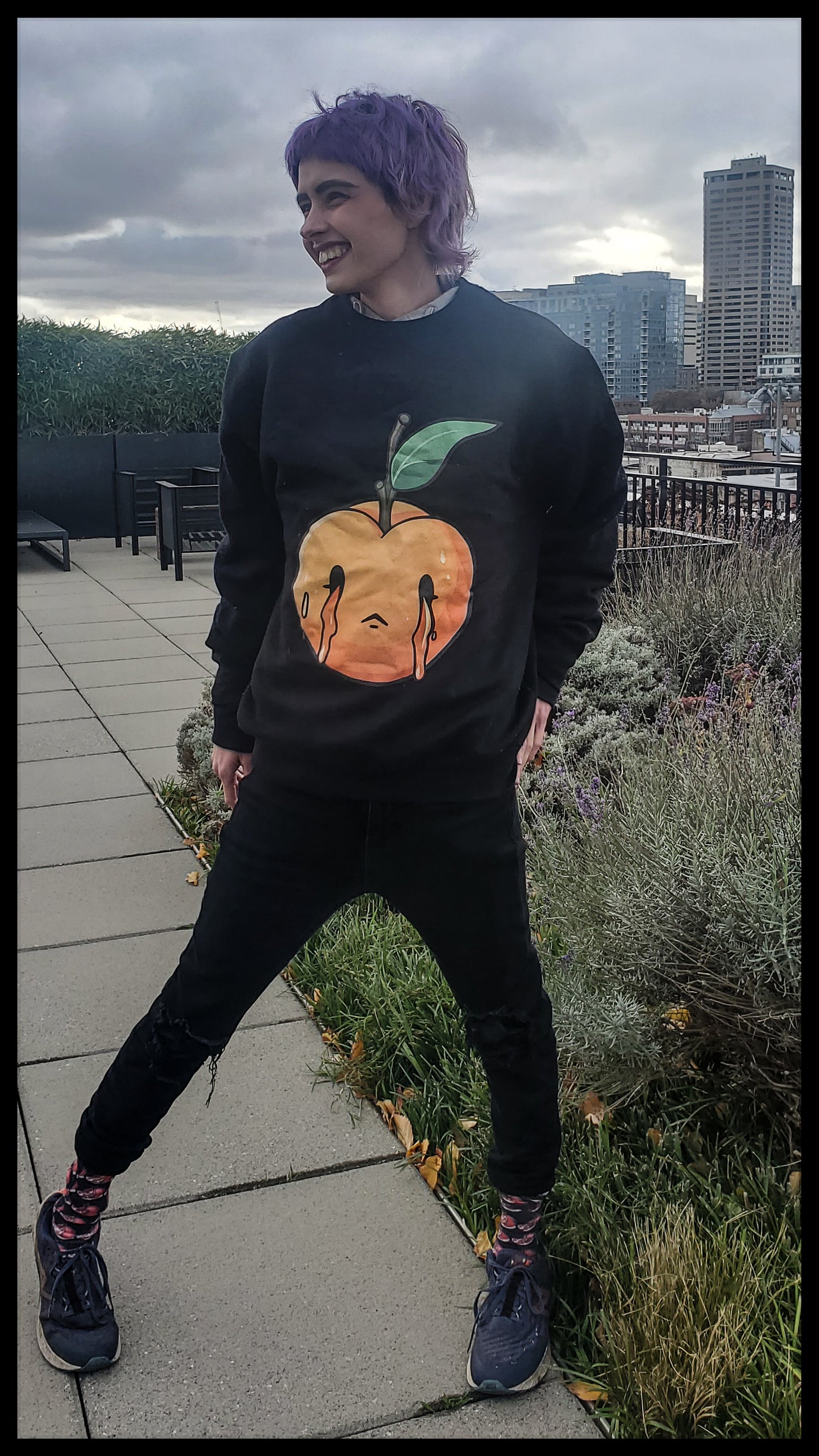 person with purple hair and a joyful expression, standing outdoors with a cityscape in the background. They are wearing a black sweatshirt featuring a graphic of a peach with black dripping lines, paired with black distressed jeans and graphic socks