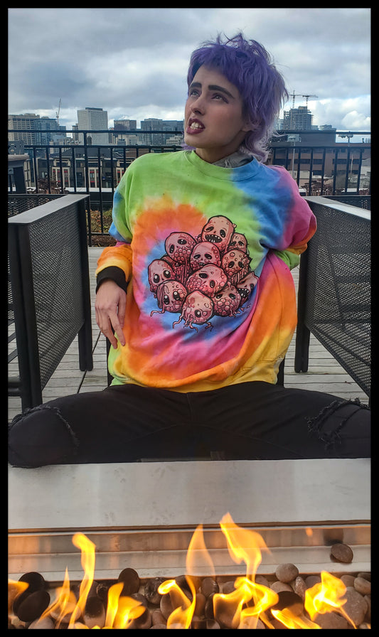 the subject is seated, sporting a vivid, tie-dyed sweatshirt with a skull motif, against an urban skyline. They have striking purple hair and a spirited expression, with a dynamic fire feature in the foreground adding warmth to the scene.