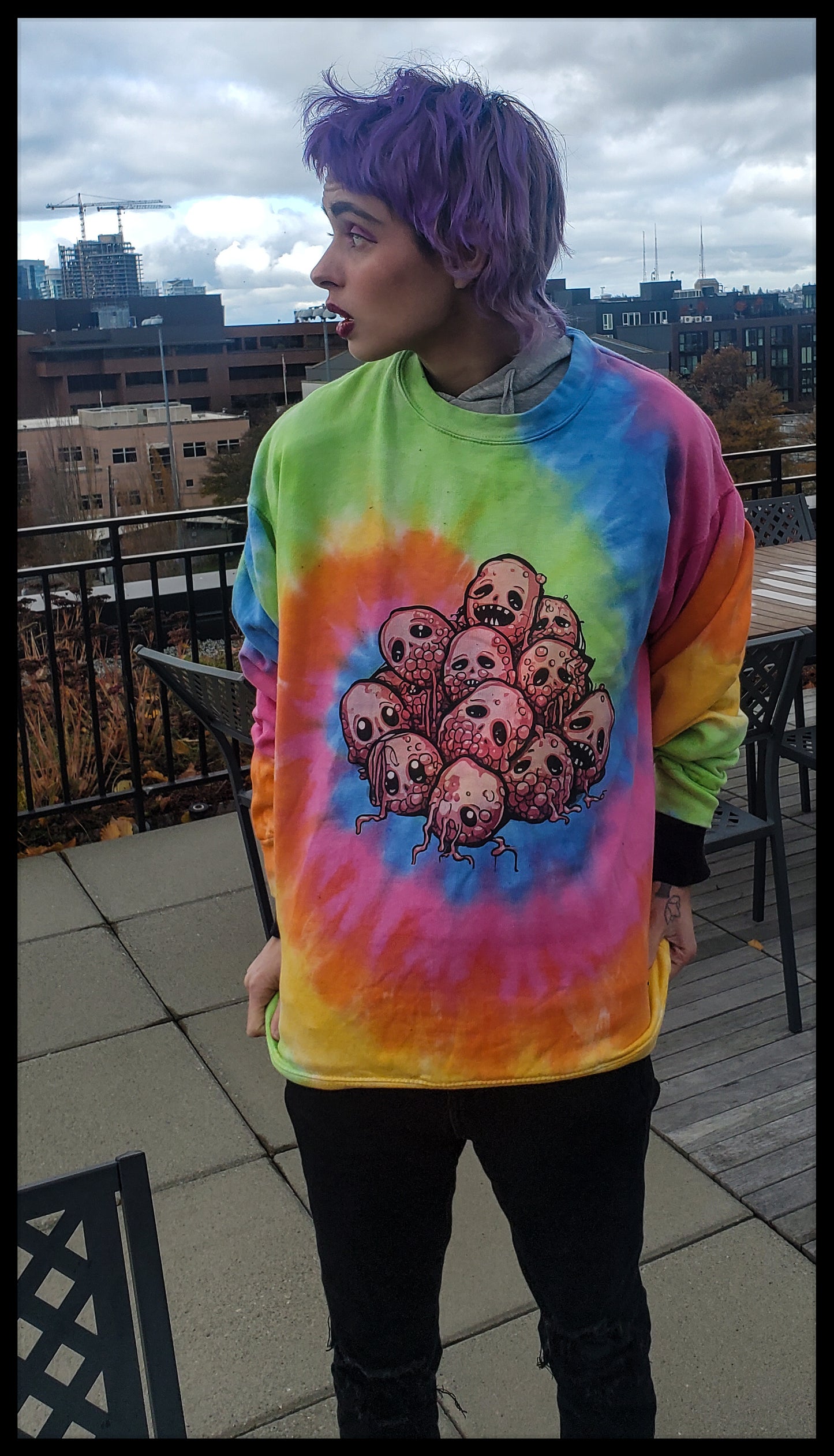 The individual is turned sideways, showcasing a vibrant tie-dye sweatshirt with a cluster of cartoonish skulls, each with a unique expression. The bold colors of the sweatshirt stand out against the urban backdrop of a city skyline. The person’s purple hair complements their playful and colorful attire, while their pose and expression suggest a lively and spirited personality.