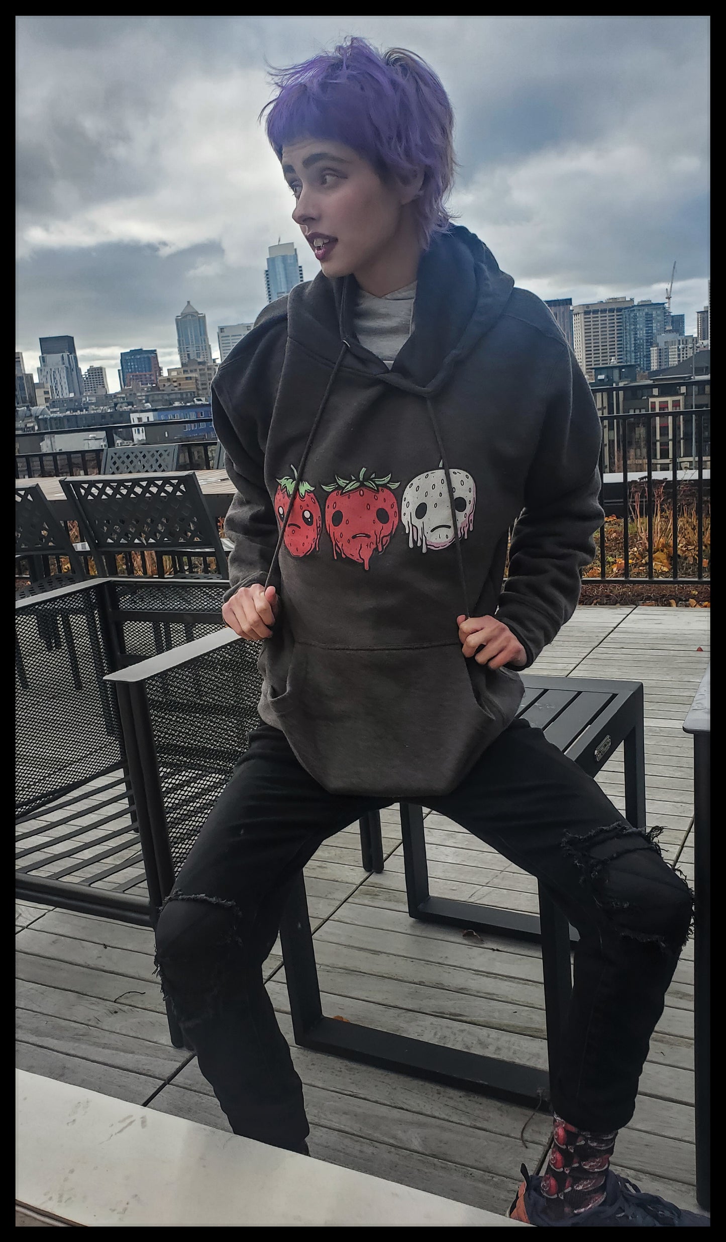 a person with purple hair and vibrant makeup. They're wearing a dark hoodie with a unique design featuring strawberries and a skull with a horror theme. The individual seems to be sitting or leaning on a chair outdoors, with an urban skyline in the background, suggesting a rooftop or balcony setting. The expression on their face is lively, and they appear to be engaged in a conversation or looking at something out of frame.