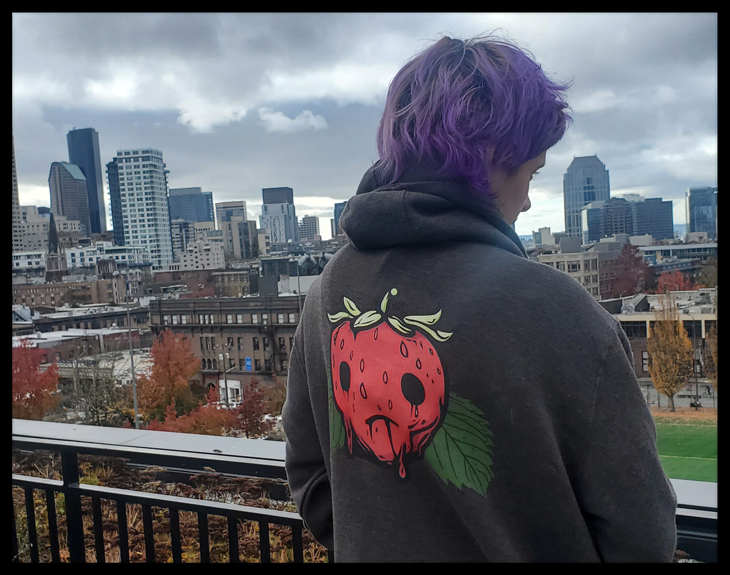 Person with purple hair wearing a hoodie with a strawberry design, standing on a rooftop with a cityscape in the background.