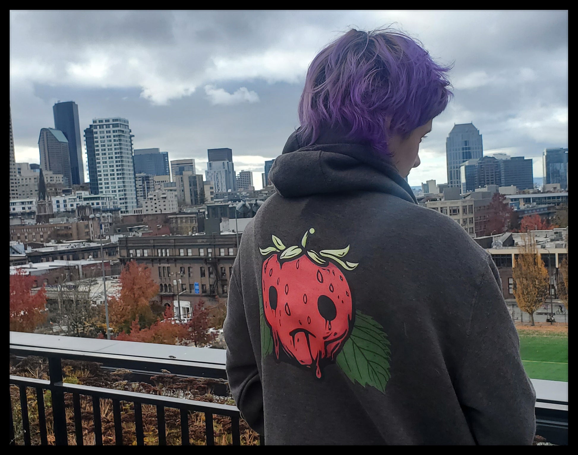 A person from the back, gazing towards a cityscape. They have purple hair and are wearing a gray hoodie featuring a design of a strawberry with dripping red juice. The individual's stance and the city skyline in the background create a contemplative scene.