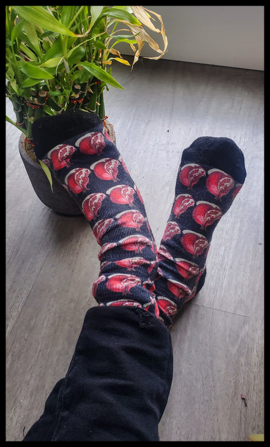 The socks display a repeated pattern of red pomegranates, with their rich, deep coloring standing out against a contrasting dark fabric, alluding to the fruit's lush and vibrant interior.