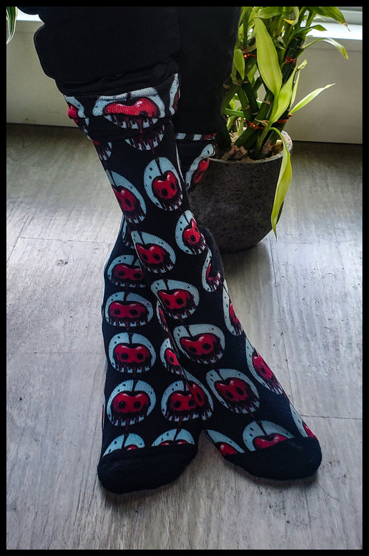 A pair of socks features a repeating cherry design with red fruit and blood-like drips, set against a dark background, worn above black pants, accentuating the striking pattern.