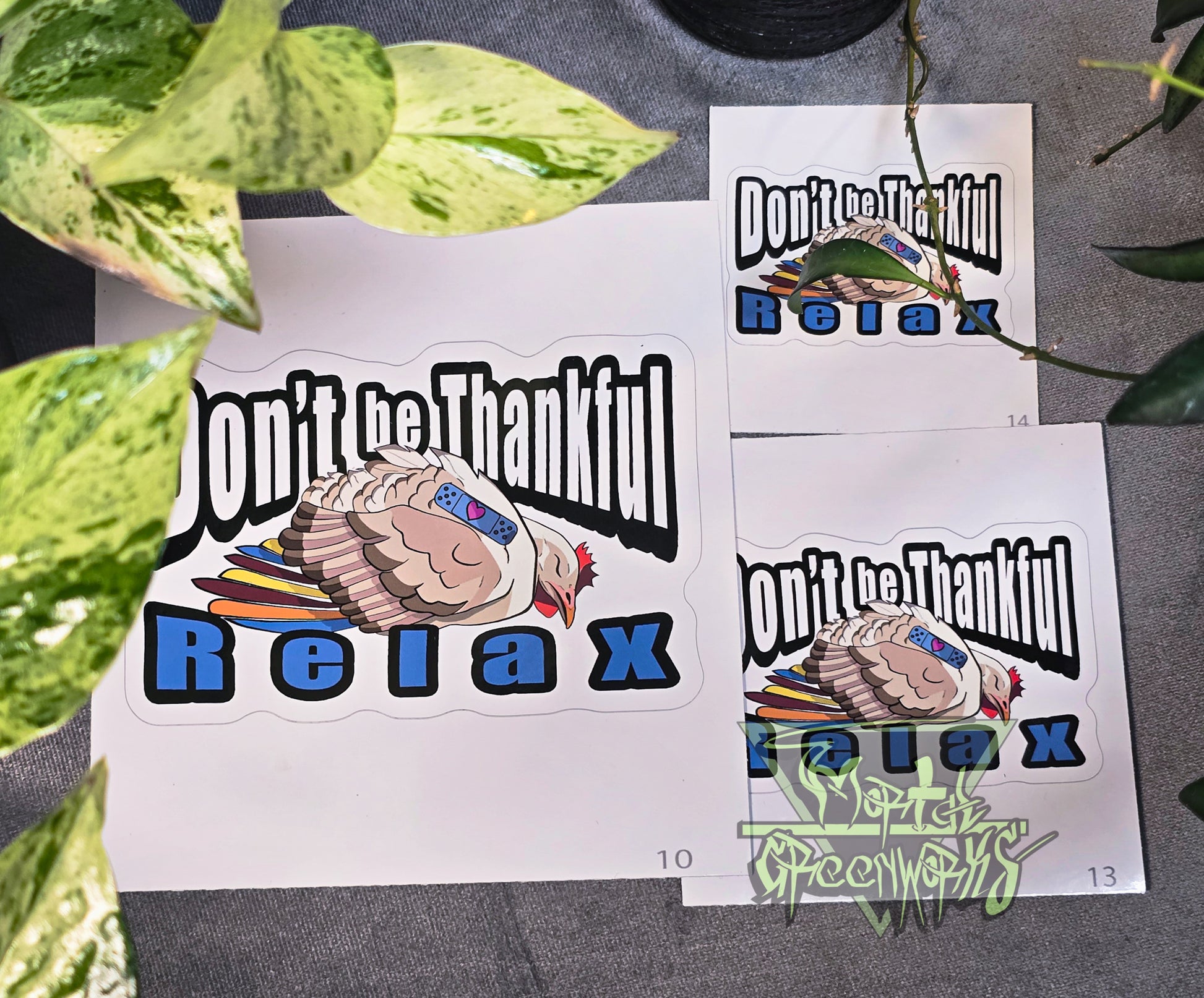 Three stickers of different sizes are on a tablecloth with a green pathos leaf peaking out of the foreground. The sticker features a beige and brown sleeping turkey with colorful feathers, and a blue sleeping Band-Aid with a heart. In bold Impact text around the turkey reads: "Don't be Thankful, Relax." The Mortal Greenworks logo appears in the bottom-right corner of the image.