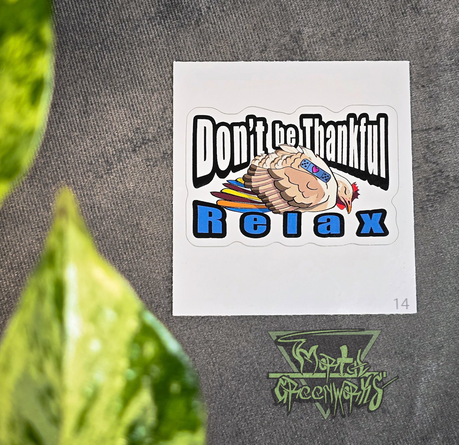 A 3x3" sticker lies on a tablecloth with a green pathos leaf peaking out of the foreground. The sticker features a beige and brown sleeping turkey with colorful feathers, and a blue sleeping Band-Aid with a heart. In bold Impact text around the turkey reads: "Don't be Thankful, Relax." The Mortal Greenworks logo sits in the bottom right of the image.