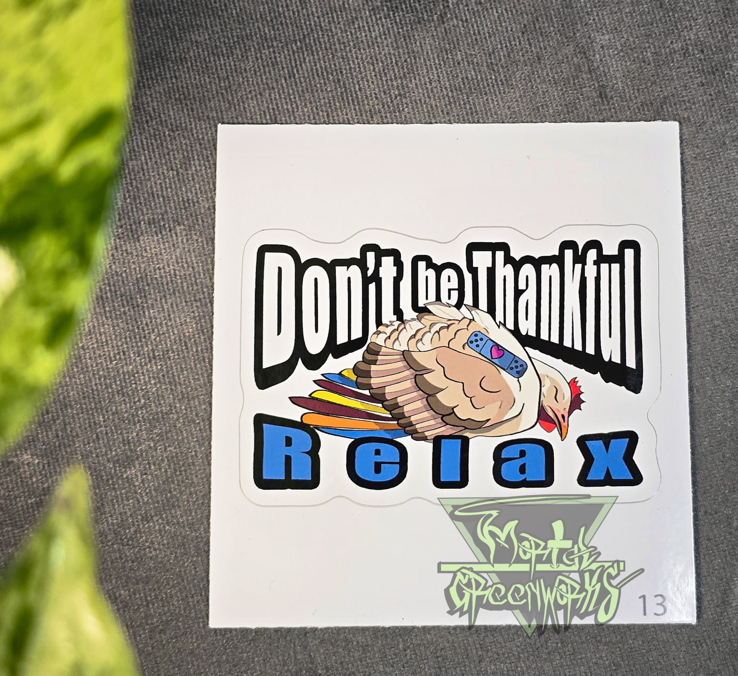 A 4x4" sticker lies on a tablecloth with a green pathos leaf peaking out of the foreground. The sticker features a beige and brown sleeping turkey with colorful feathers, and a blue sleeping Band-Aid with a heart. In bold Impact text around the turkey reads: "Don't be Thankful, Relax." The Mortal Greenworks logo sits in the bottom right of the image.