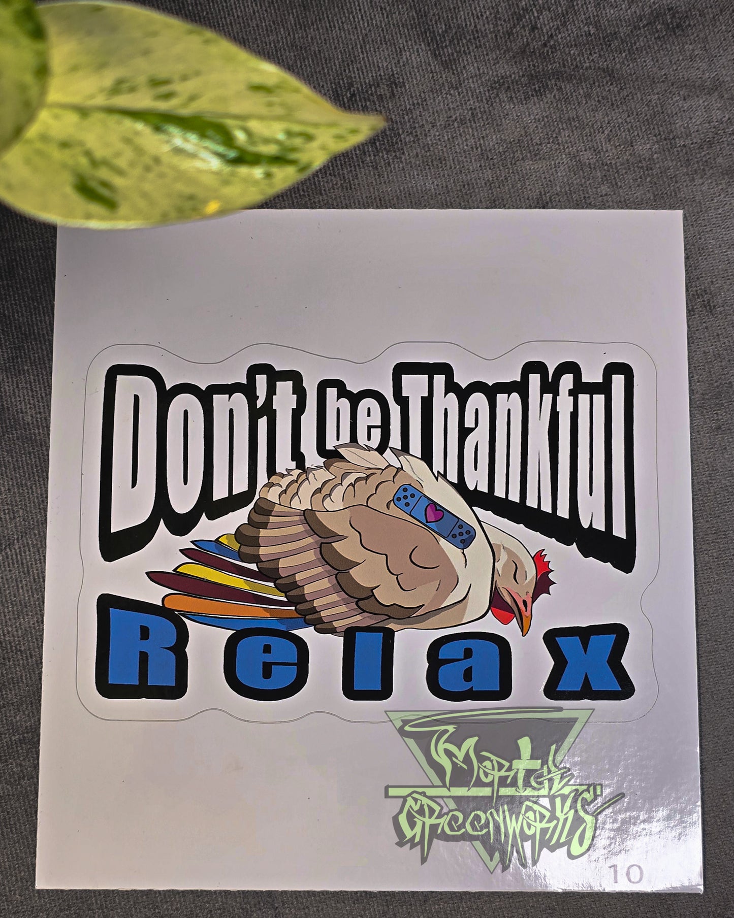 A 6x6" sticker lies on a tablecloth with a green pathos leaf peaking out of the foreground. The sticker features a beige and brown sleeping turkey with colorful feathers, and a blue sleeping Band-Aid with a heart. In bold Impact text around the turkey reads: "Don't be Thankful, Relax." The Mortal Greenworks logo sits in the bottom right of the image.