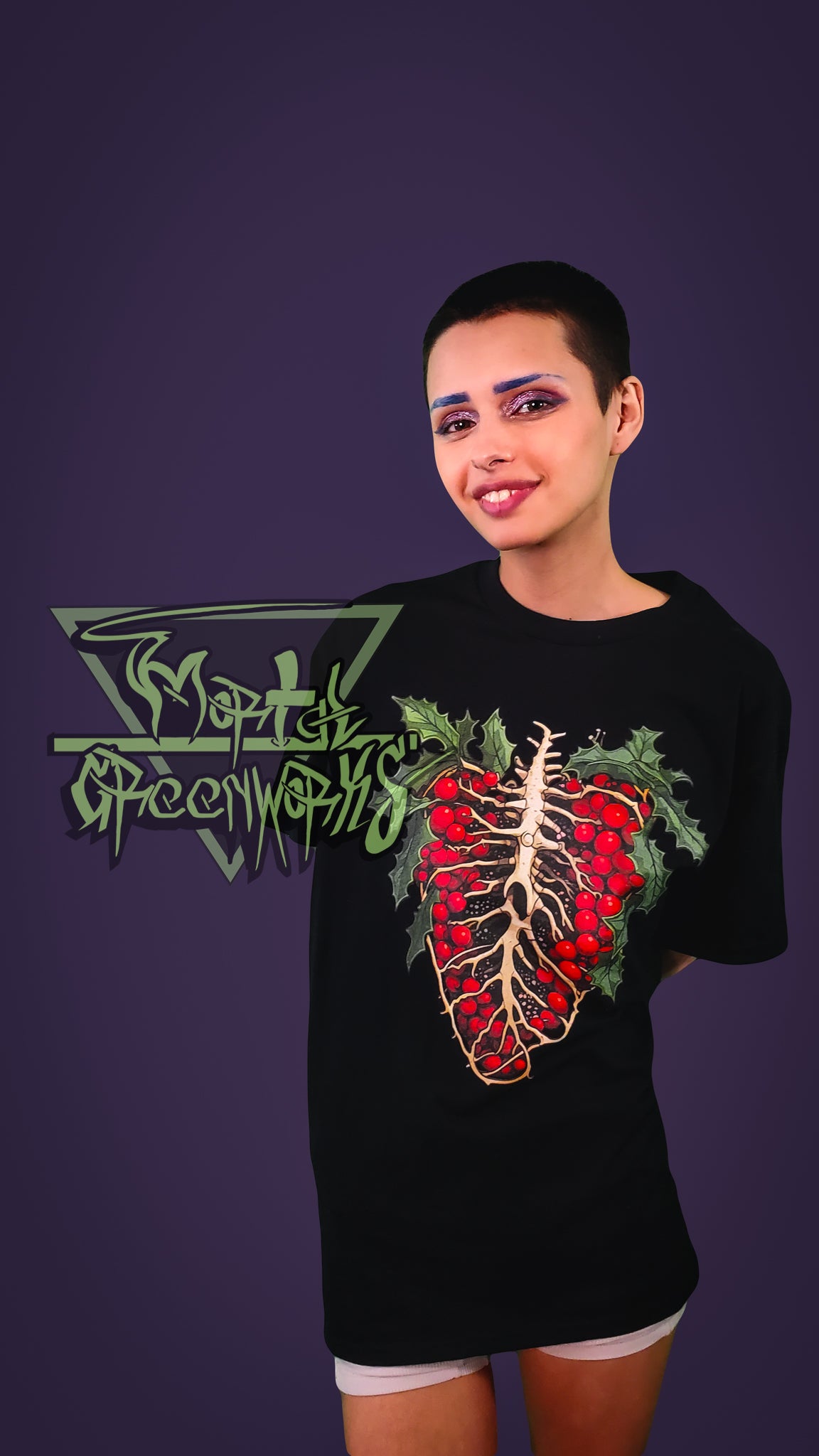 A smiling Person wearing a black t-shirt with a red and green design of a skeletal ribcage with holly growing around it on a purple background
