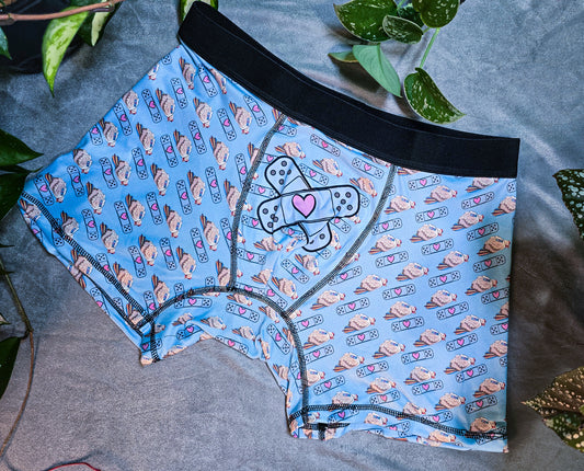 Boxer briefs in light blue featuring a whimsical pattern of chickens and bandaids with hearts, laid flat on a gray surface, surrounded by lush green plants for a vibrant contrast.