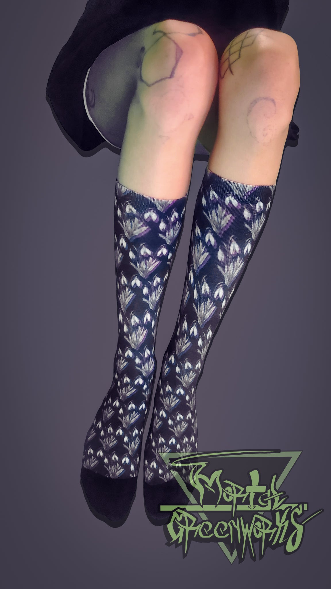 A model with tattoos on their knees is laying down and displaying the black, purple, white and green Snowdrop Flower socks on black cotton. The image background is light grey and the Mortal Greenworks Logo displays on the right hand side at the bottom.