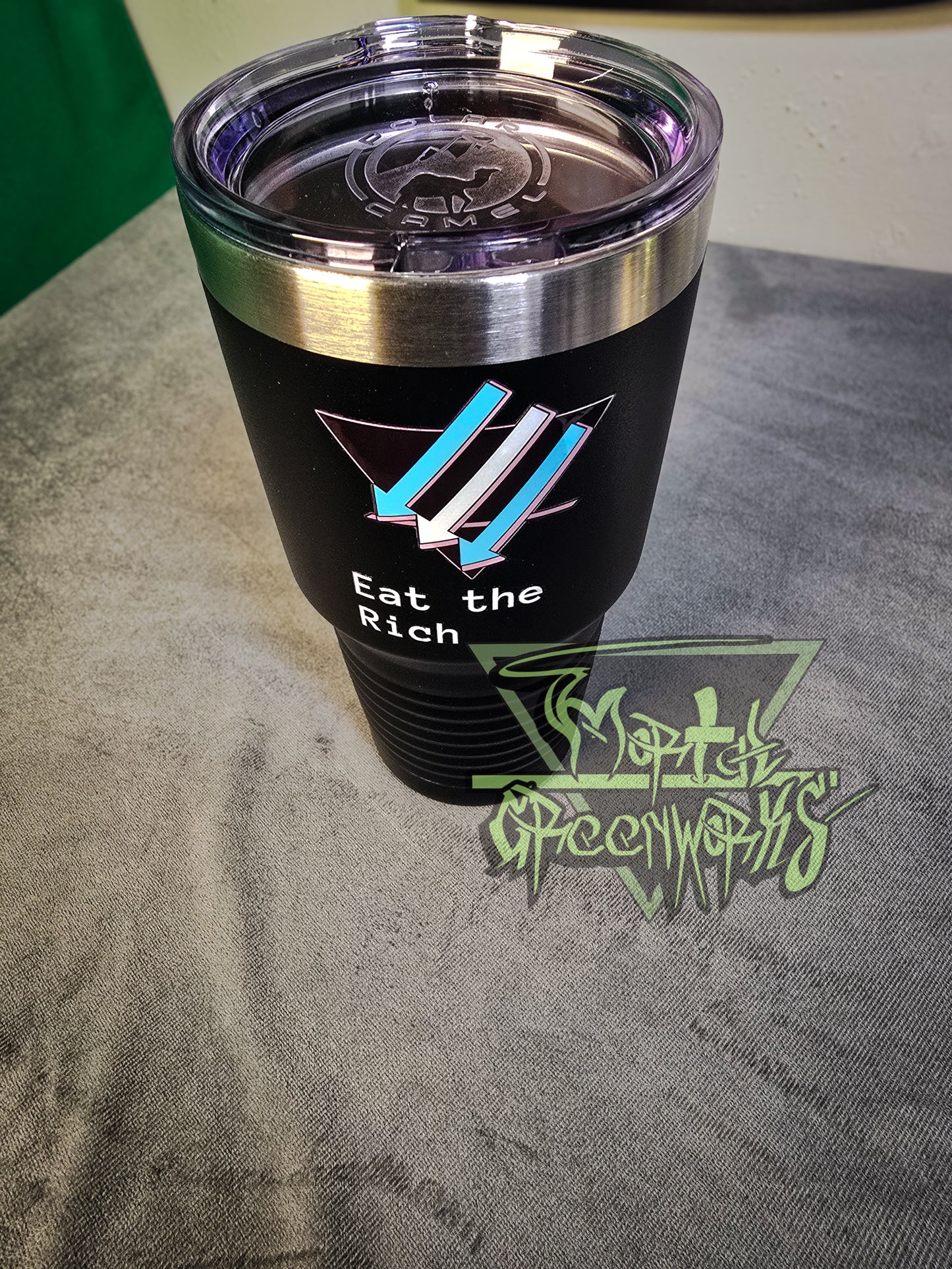 A close up photograph of tall, steel black Tumbler travel mug with a chrome rim at the top and the climate front trans pride flag stylization on the side. A plastic lid with a mouth piece sits on top. The custom text below the logo says eat the rich. The mug sits on a grey table cloth with a green and white wall peeking out in the foreground.