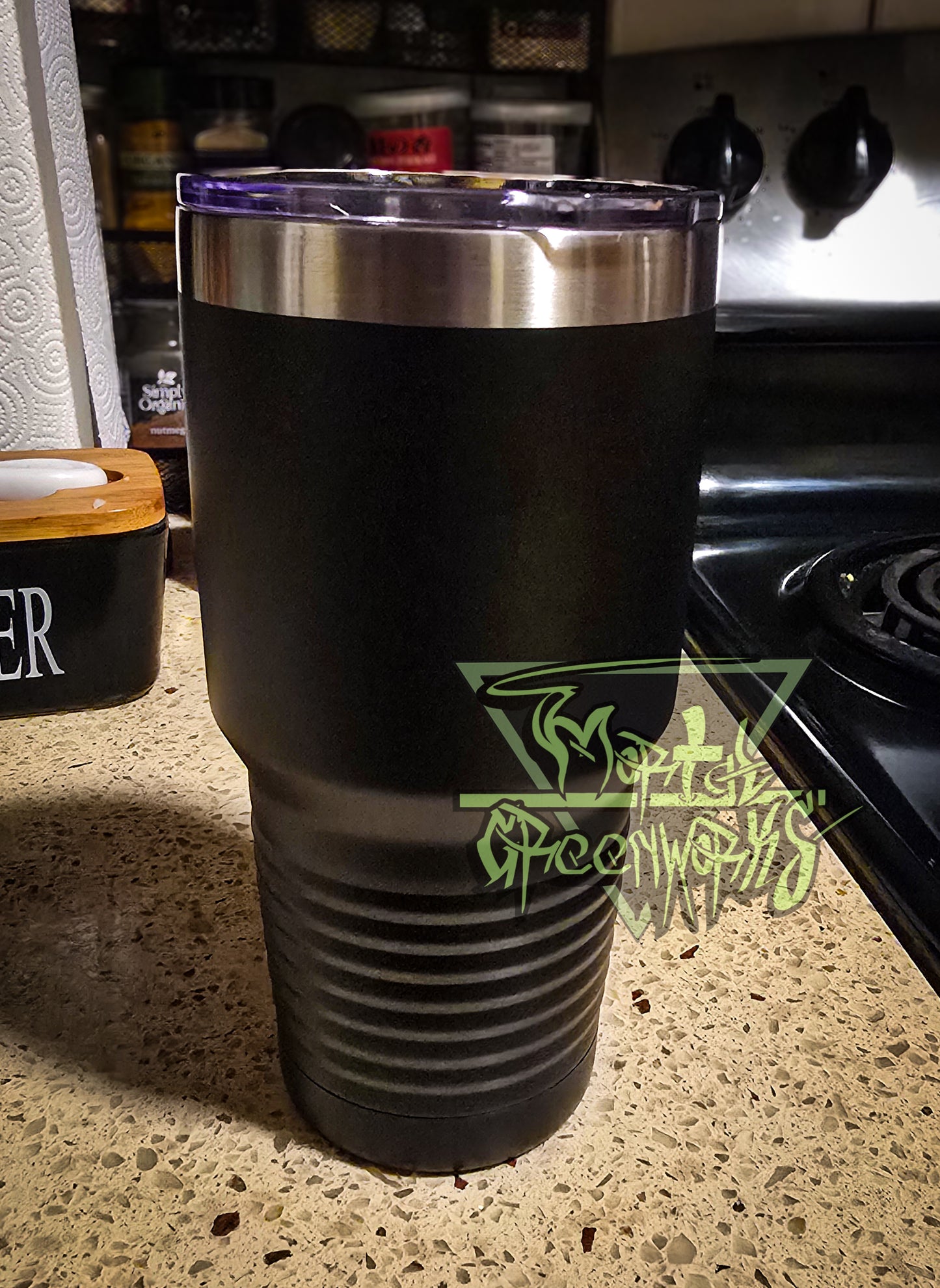 A close-up photograph of a tall, steel black Tumbler travel mug with a plastic lid with a mouthpiece sits on top. The mug sits on a granite countertop with a butter box, paper towels, and spices in the background. A transparent Mortal Greenwors watermark lingers to the right side of the image.
