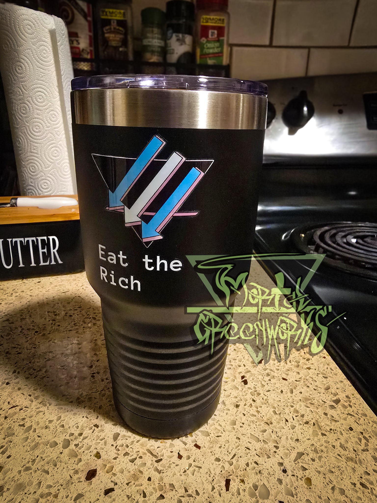 A close up photograph of tall, steel black Tumbler travel mug with a chrome rim at the top and the climate front trans pride flag stylization on the side. A plastic lid with a mouth piece sits on top. The custom text below the logo says eat the rich. The mug sits on a granite countertop with a butter box, paper towels, and spices in the background.