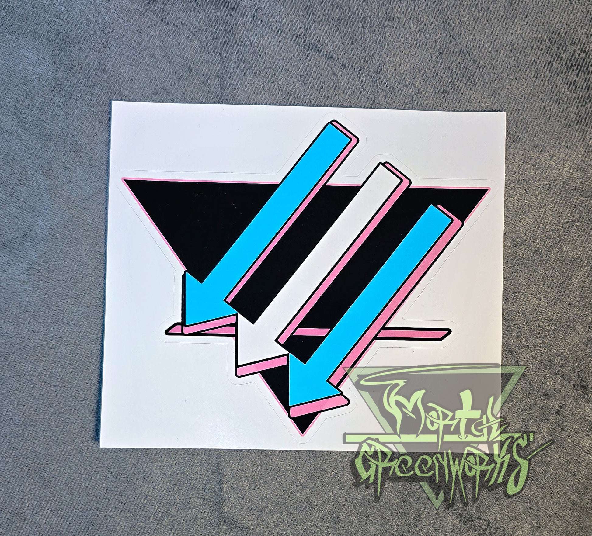 A close-up photograph of a  4x4 sticker placed on a light grey tablecloth. The design is a black upside-down triangle with a pink line in the bottom third, with light blue and white arrows pointing left and downward. The Mortal Greenworks Watermark is in the bottom right-hand corner of the image.