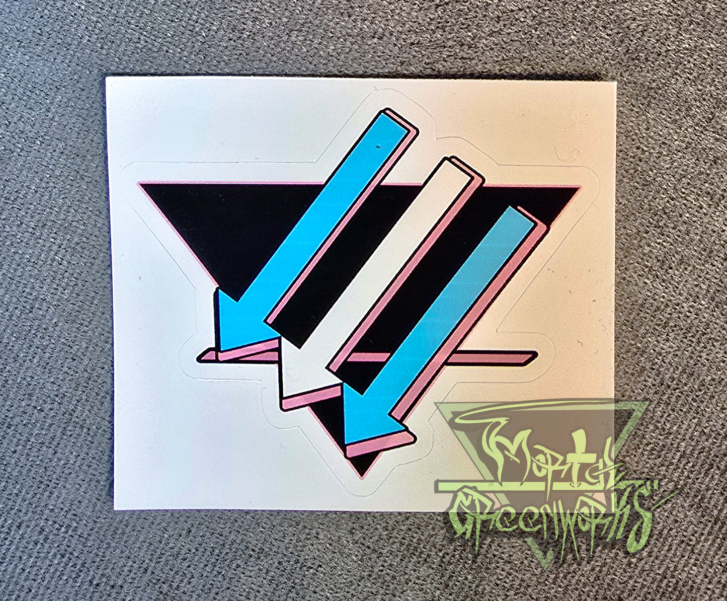 A close up photograph of a  3x3 sticker placed on a light grey tablecloth. The design is a black upside down triangle with a pink line in the bottom third with light blue and white arrows pointing left and downward. The Mortal Greenworks Watermark is in the bottom right hand corner of the image.