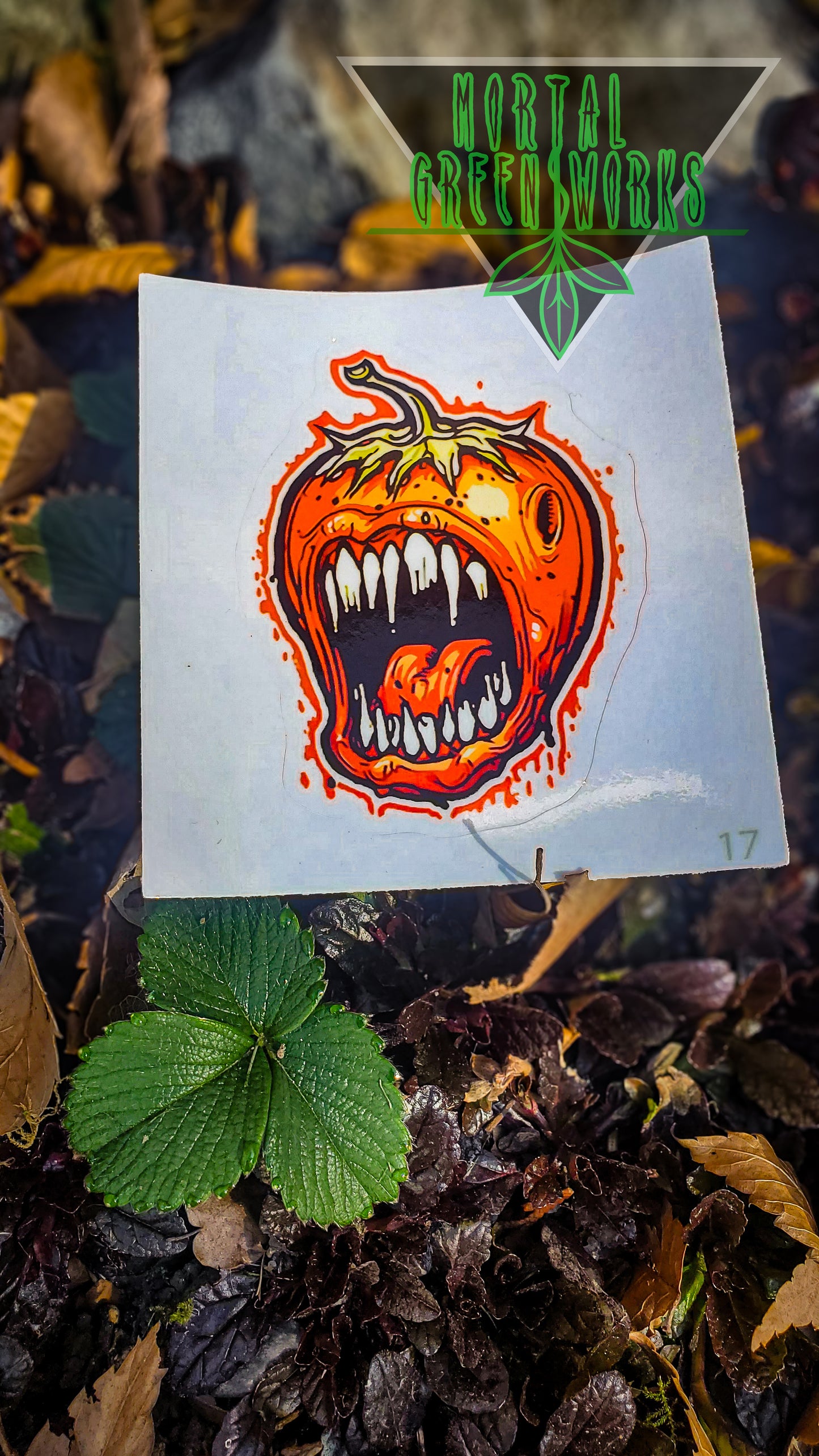 A menacing strawberry creature sticker against a backdrop of fallen leaves and the 'Mortal Greenworks' logo.