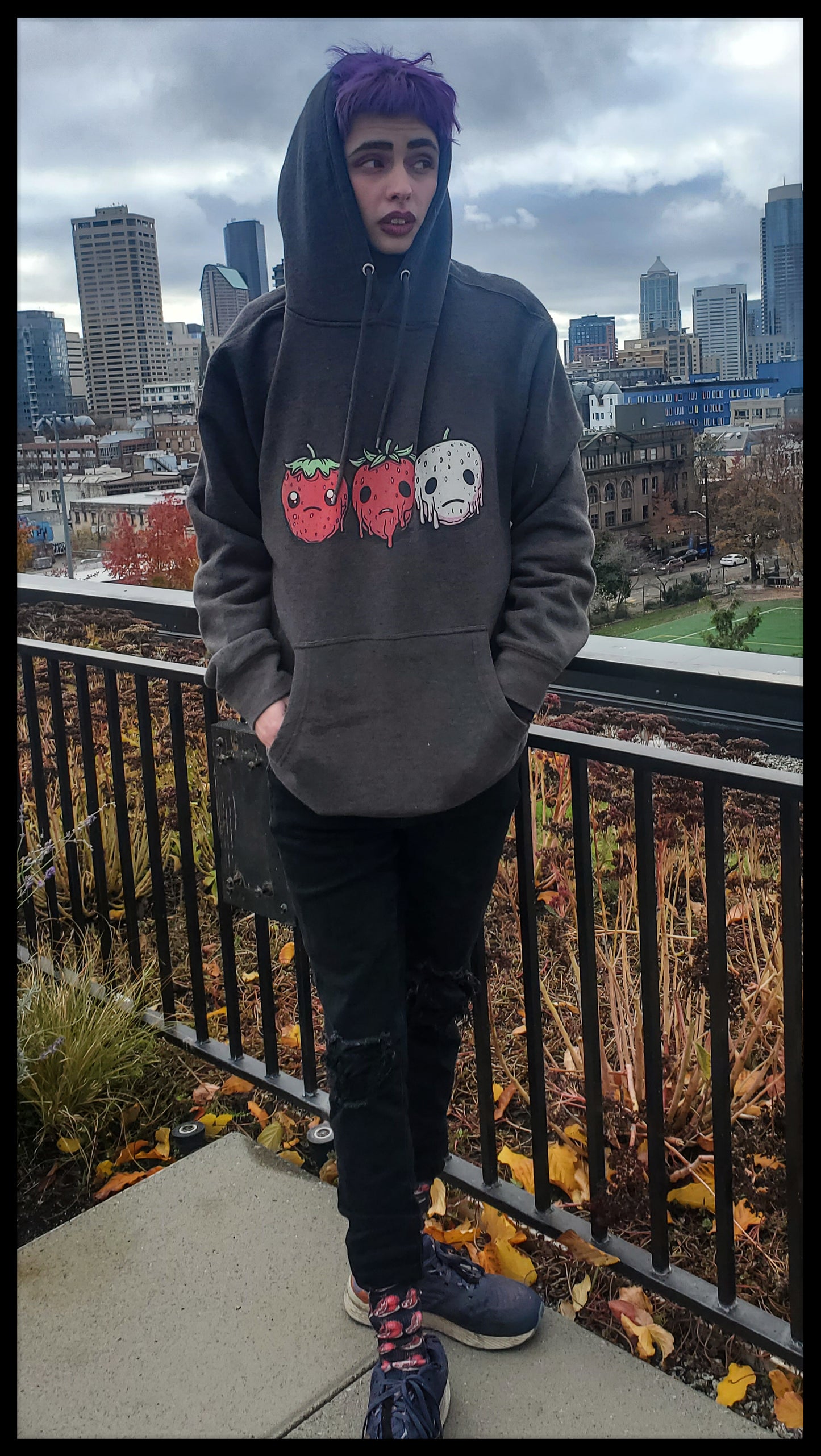 A Person with purple hair is wearing a hoodie on an urban balcony, with three smiling strawberries: one white and the other two red.