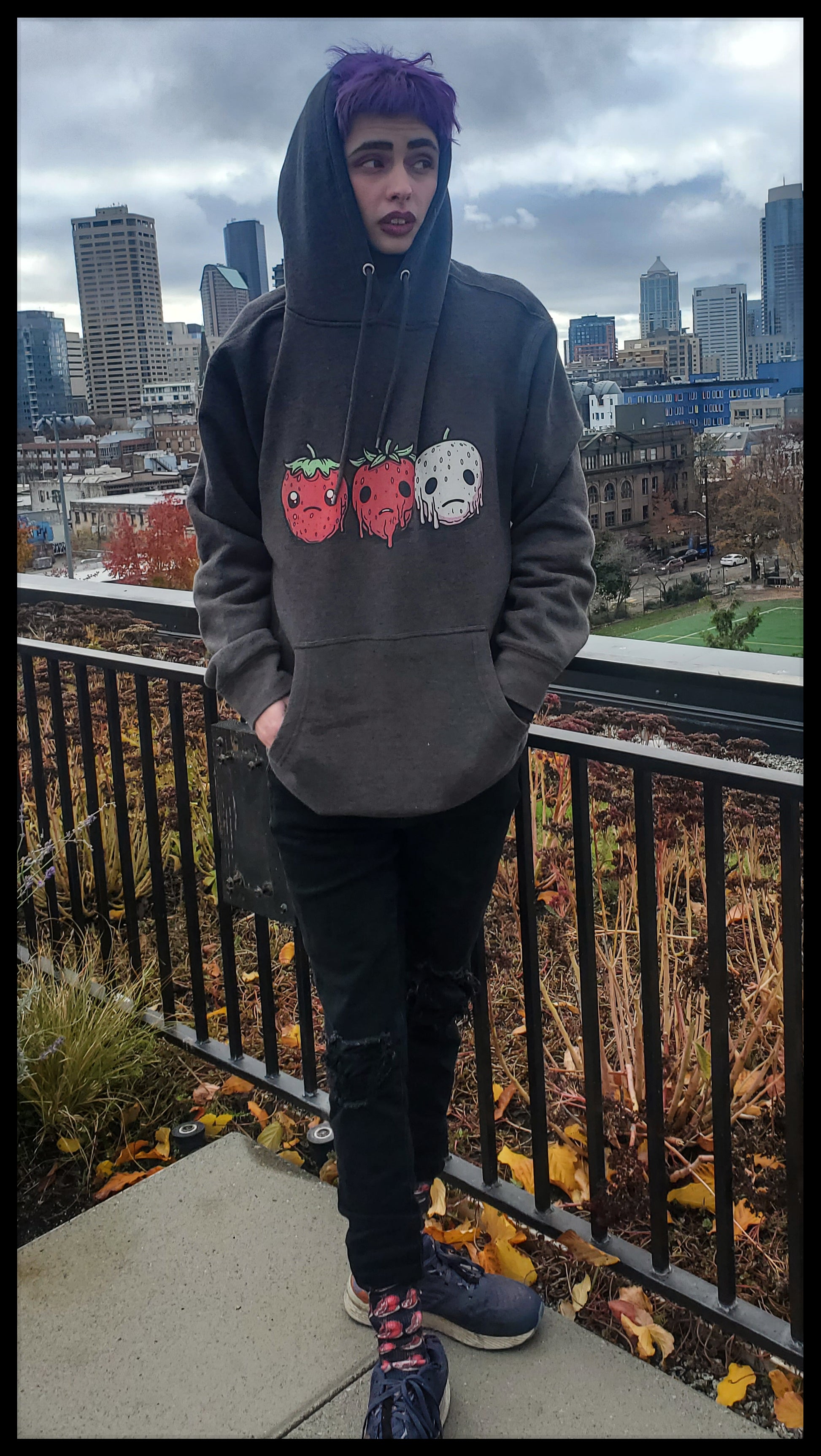 A Person with purple hair is wearing a hoodie on an urban balcony, with three smiling strawberries: one white and the other two red.