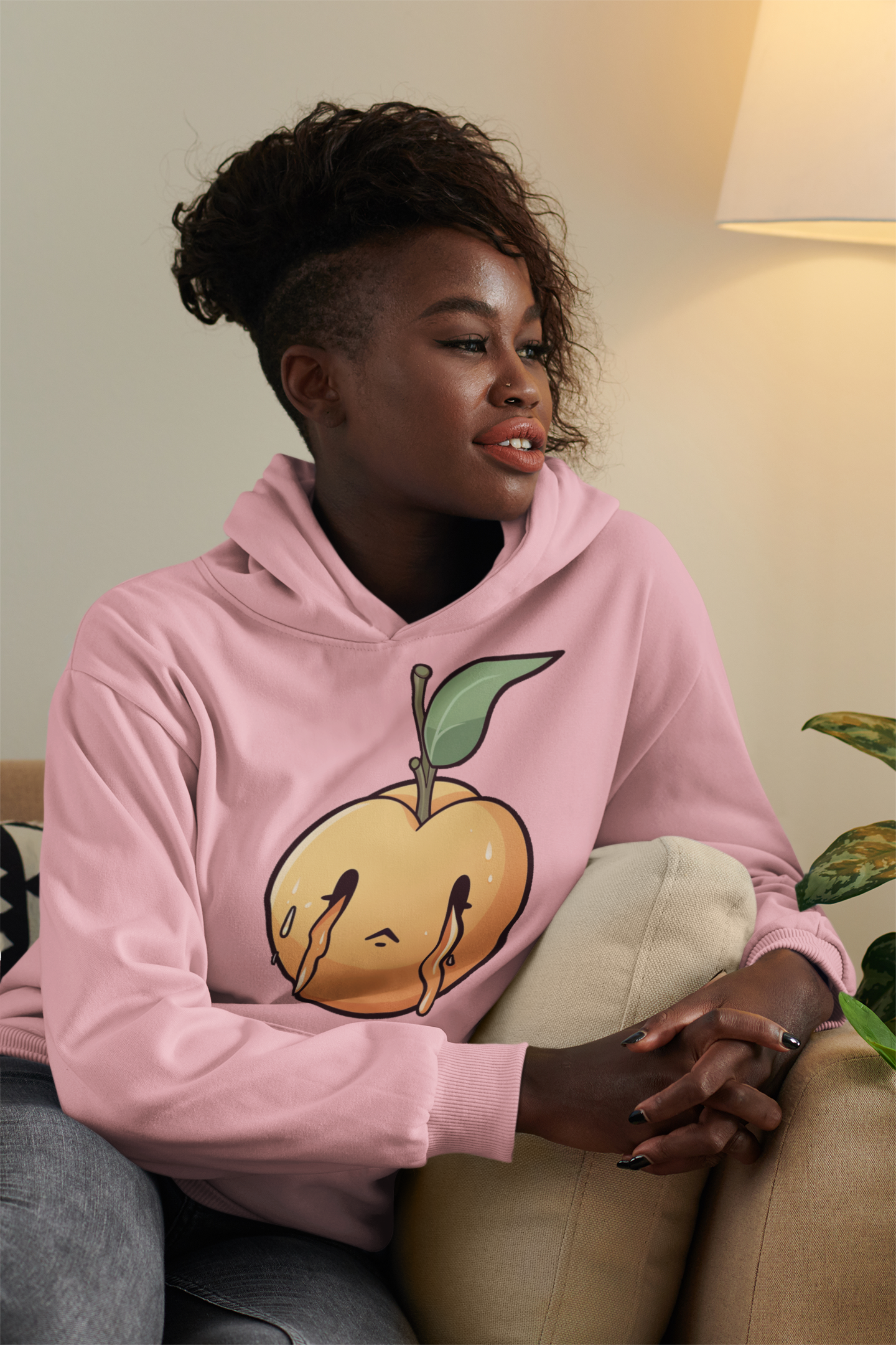 Woman sitting on a couch, smiling, wearing a pink hoodie with a melting peach design, indoor setting.