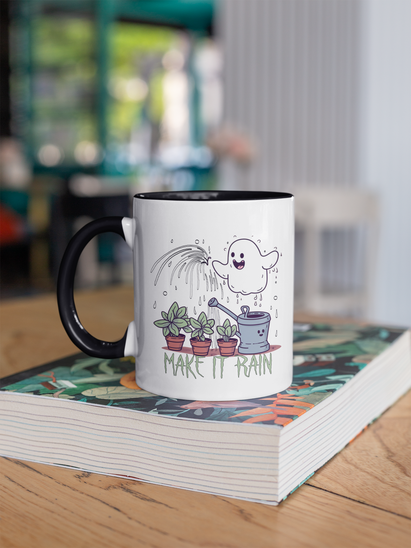 coffee mug with a creative design. The mug has a white exterior with a black handle, and on its surface, there is an illustration of a joyful ghost watering three potted plants with a water can. Above the scene, the words "MAKE IT RAIN" are written, adding a playful touch to the design. The mug is situated on a book with a colorful cover that rests on a wooden table, and the background shows an indoor setting with a view of greenery outside a window. The overall impression is one of che