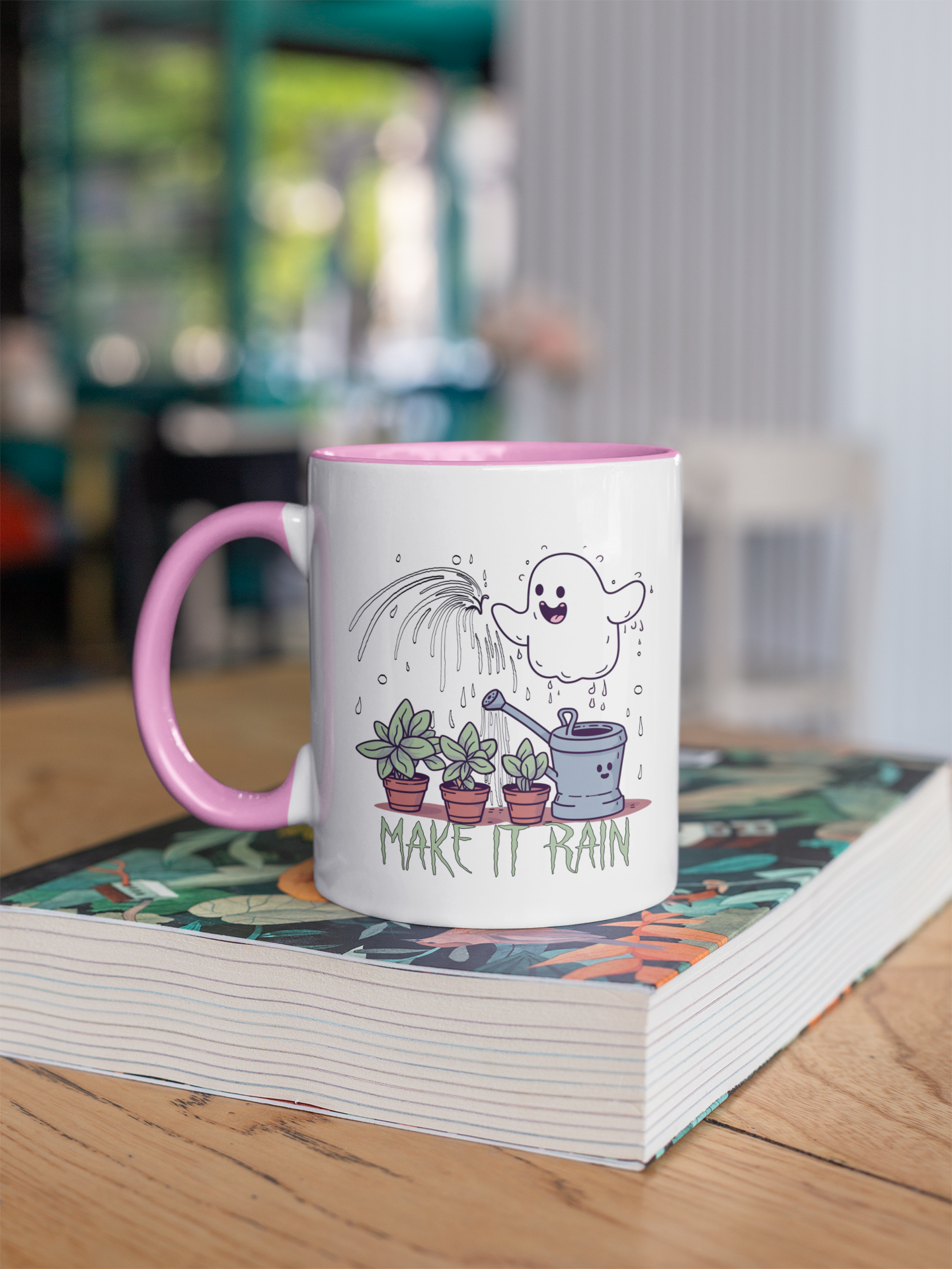 This mug features a white exterior with a pink handle and interior. The design on the mug showcases a cheerful ghost watering potted plants under a shower of rain with the phrase "MAKE IT RAIN" beneath the illustration. The pots contain green plants, and the watering can is also depicted in green. The artwork presents a lighthearted take on plant care, with a whimsical ghost adding a fun twist to the gardening theme.