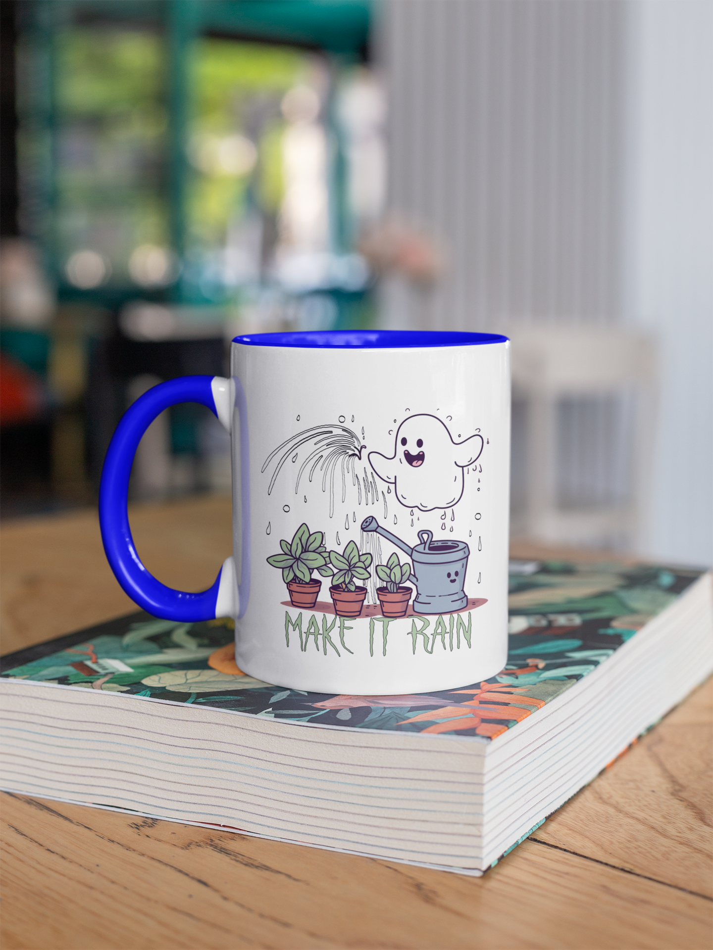 two-toned coffee mug with a white body and a blue handle. On the mug, there is a playful illustration featuring a ghost character watering three potted plants with a phrase "MAKE IT RAIN" in capital letters below the scene. The ghost appears happy, depicted with a smiling face, and it's watering the plants with a rain cloud. The design conveys a whimsical and light-hearted vibe, blending themes of nature care with a touch of humor. The mug is placed on a book with a floral cover design, wh